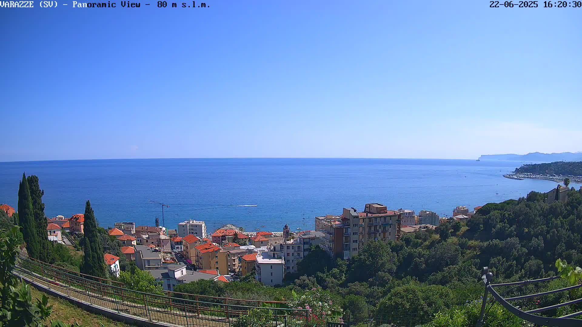 A coastal town with red-roofed buildings sits on a hillside overlooking a calm, blue sea under a clear, sunny sky.