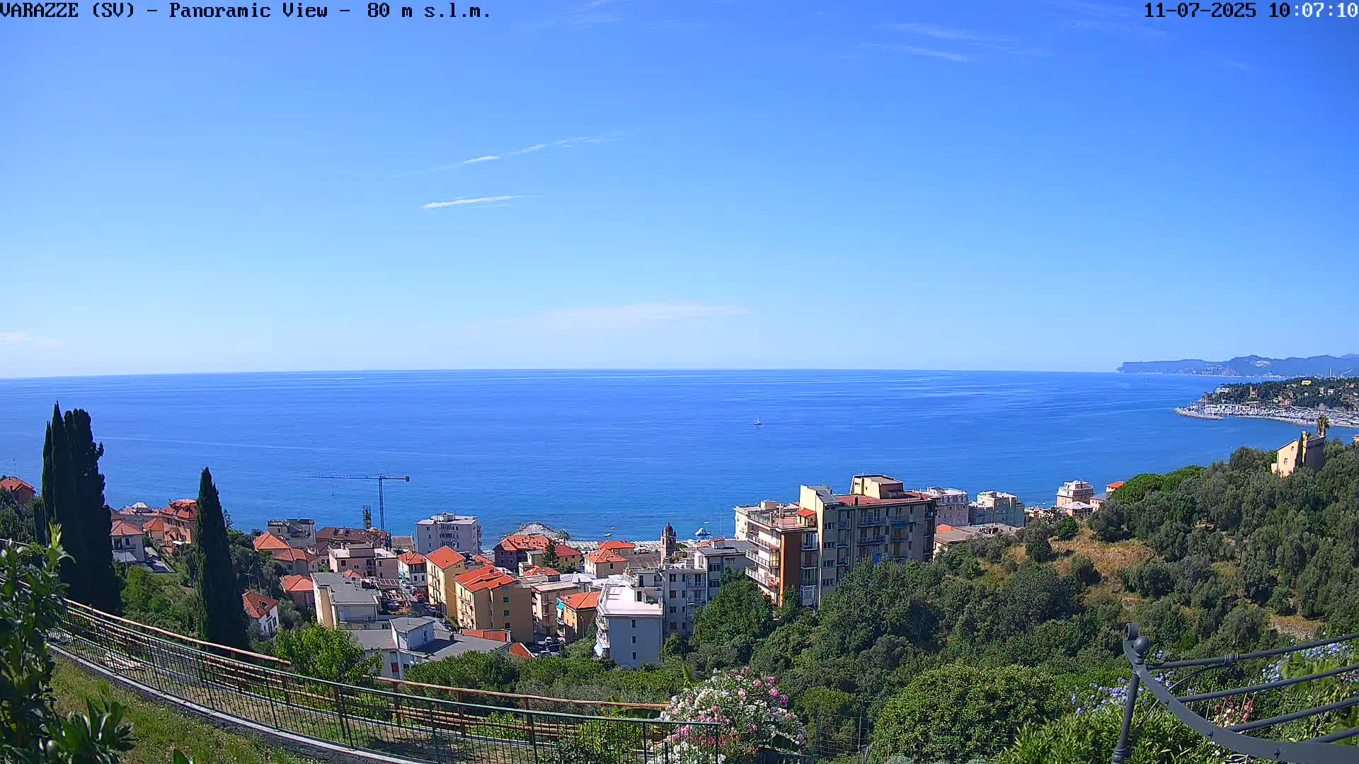 A coastal town with red-roofed buildings nestled among green hills overlooks a calm, blue ocean under a clear, sunny sky.