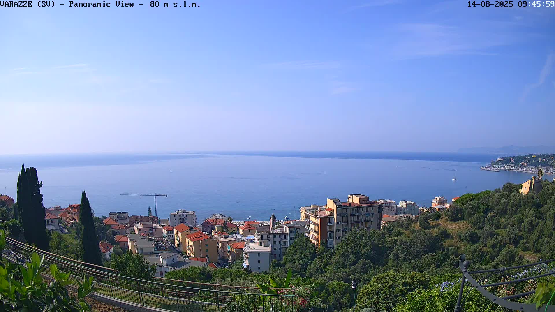 A panoramic view shows a coastal town nestled among green hills overlooking a calm, blue sea under a clear, sunny sky.