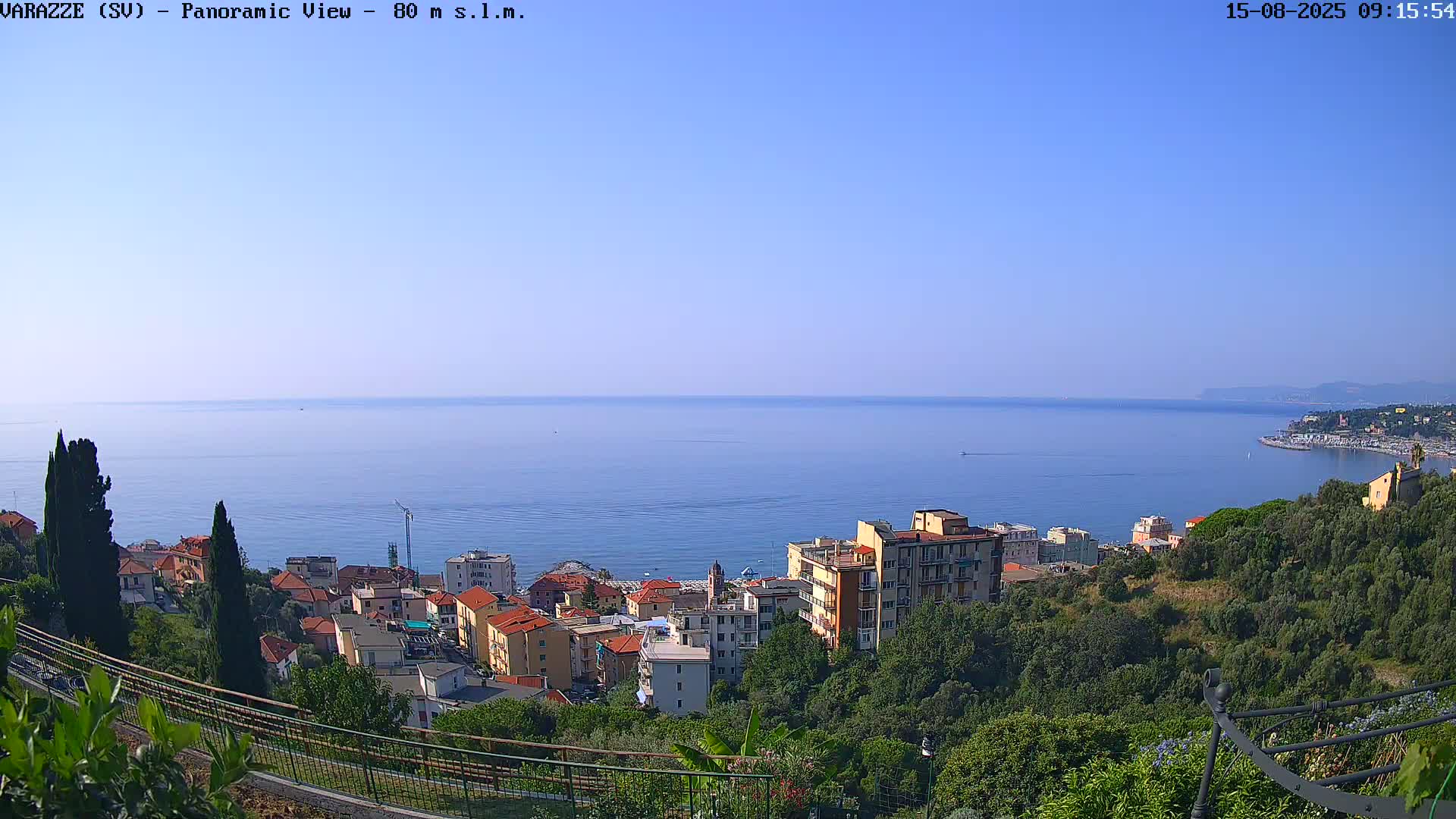 A panoramic view shows a coastal town nestled on a hillside overlooking a calm, blue sea under a clear sky.
