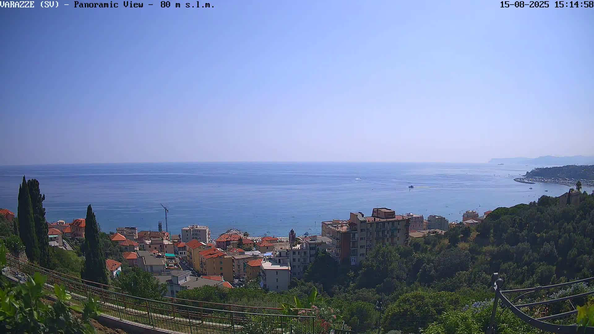 A panoramic view shows a coastal town nestled on a hillside overlooking a calm, blue sea under a clear sky.