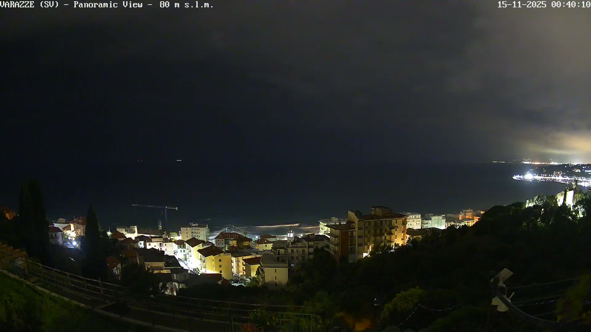 A panoramic nighttime view reveals a brightly lit coastal town nestled along a dark, wavy sea, with distant city lights illuminating a mostly cloudy sky.