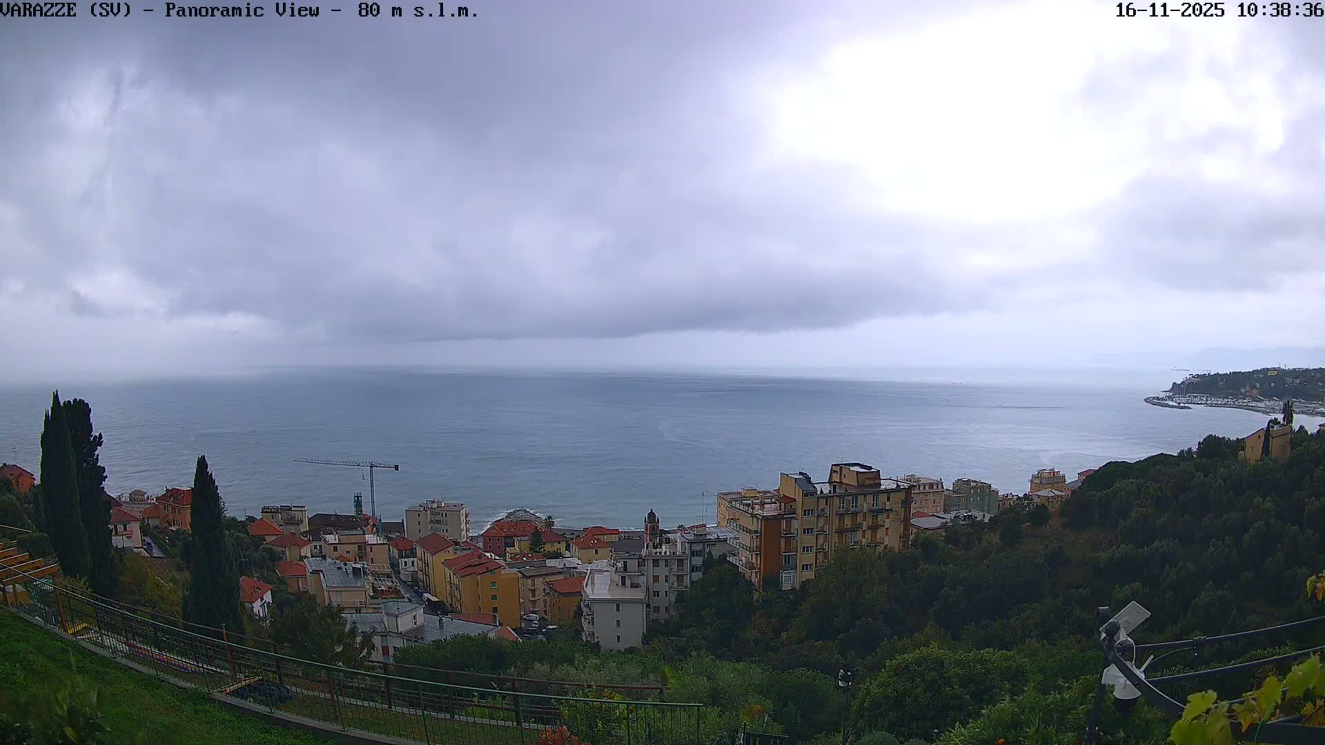 A panoramic view showcases a coastal town with colorful buildings nestled along green hillsides overlooking a calm, blue-grey sea under a heavily overcast and cloudy sky.