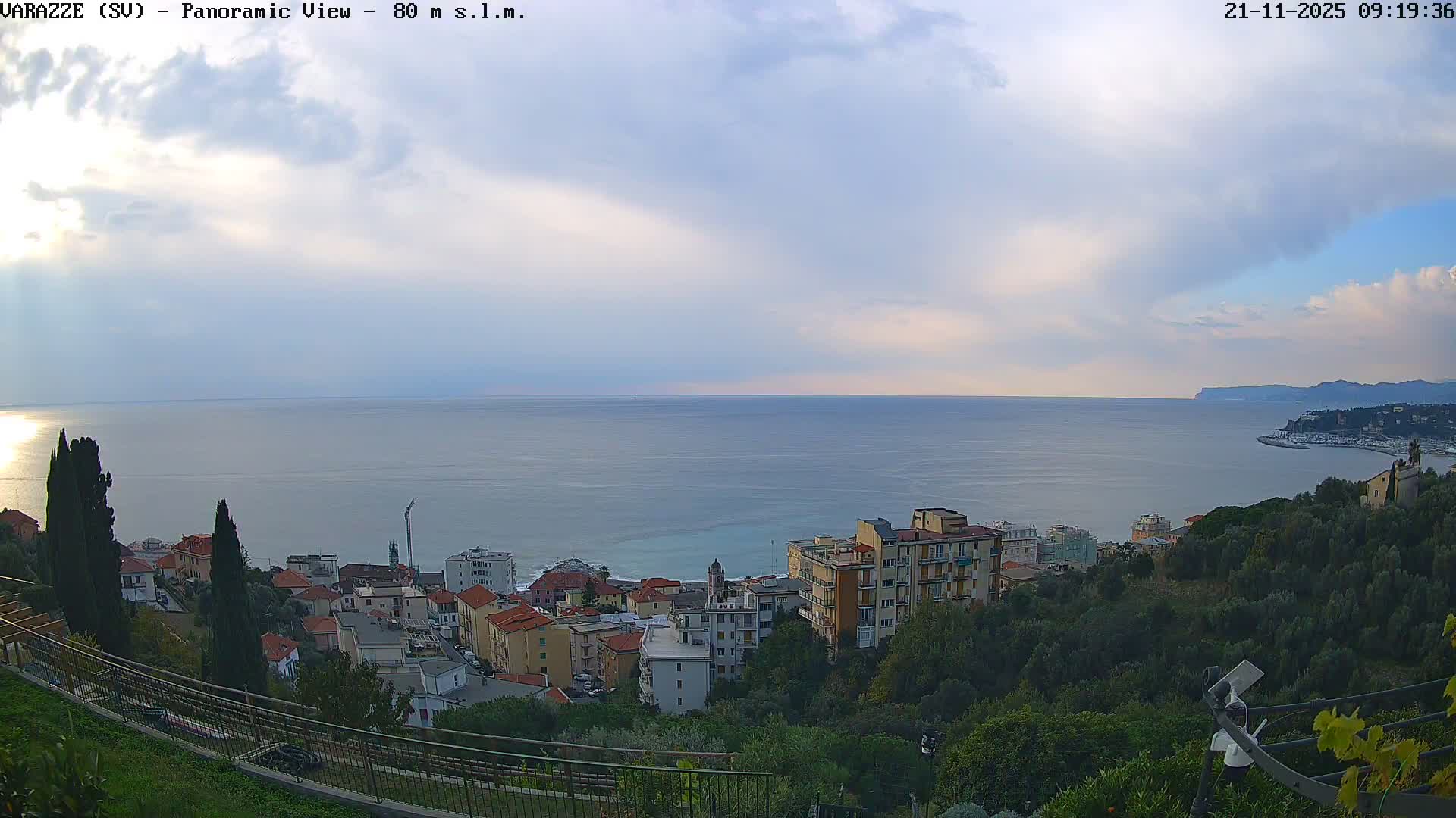 A panoramic view shows a coastal town with buildings nestled among green hills overlooking a calm sea and a distant marina, all under a partly cloudy sky with bright sunlight reflecting on the water.