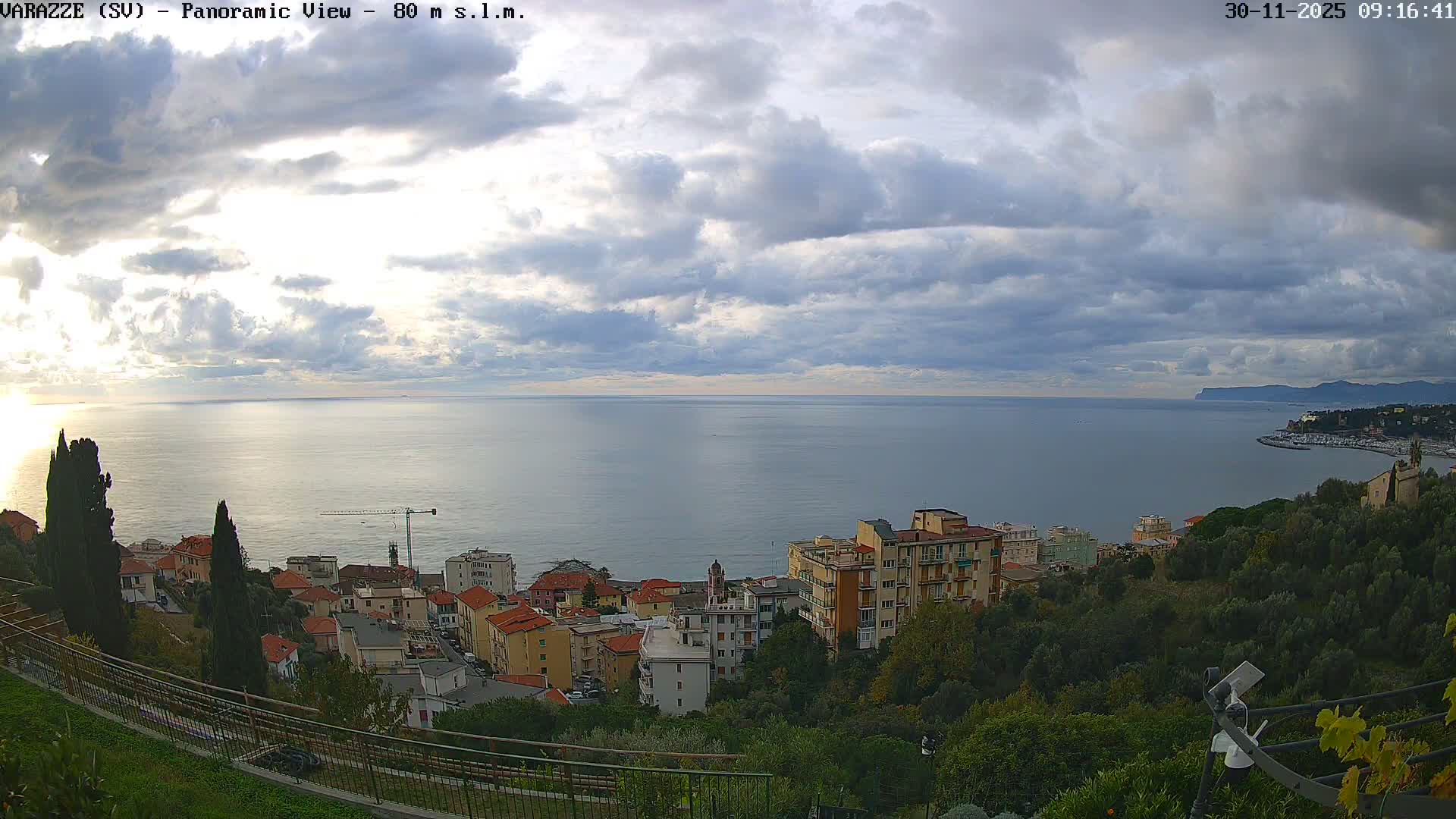 A panoramic view captures a coastal town nestled on a hillside beside a calm sea, under a partly cloudy sky with bright patches of sunlight breaking through.