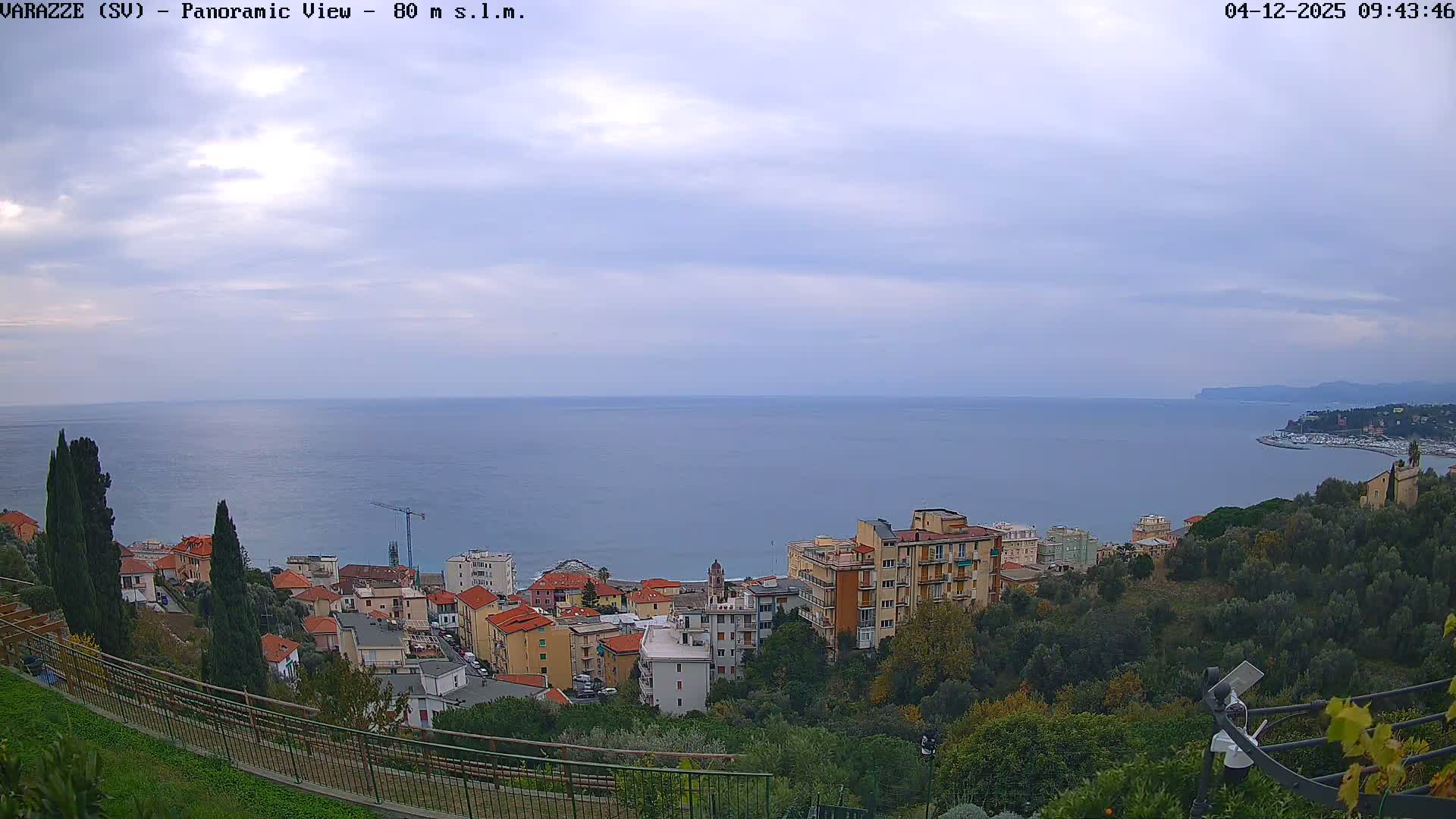 A panoramic view reveals a vibrant coastal town nestled along a calm blue sea and green hills, all under an overcast sky, with a distant curving shoreline leading to a marina.