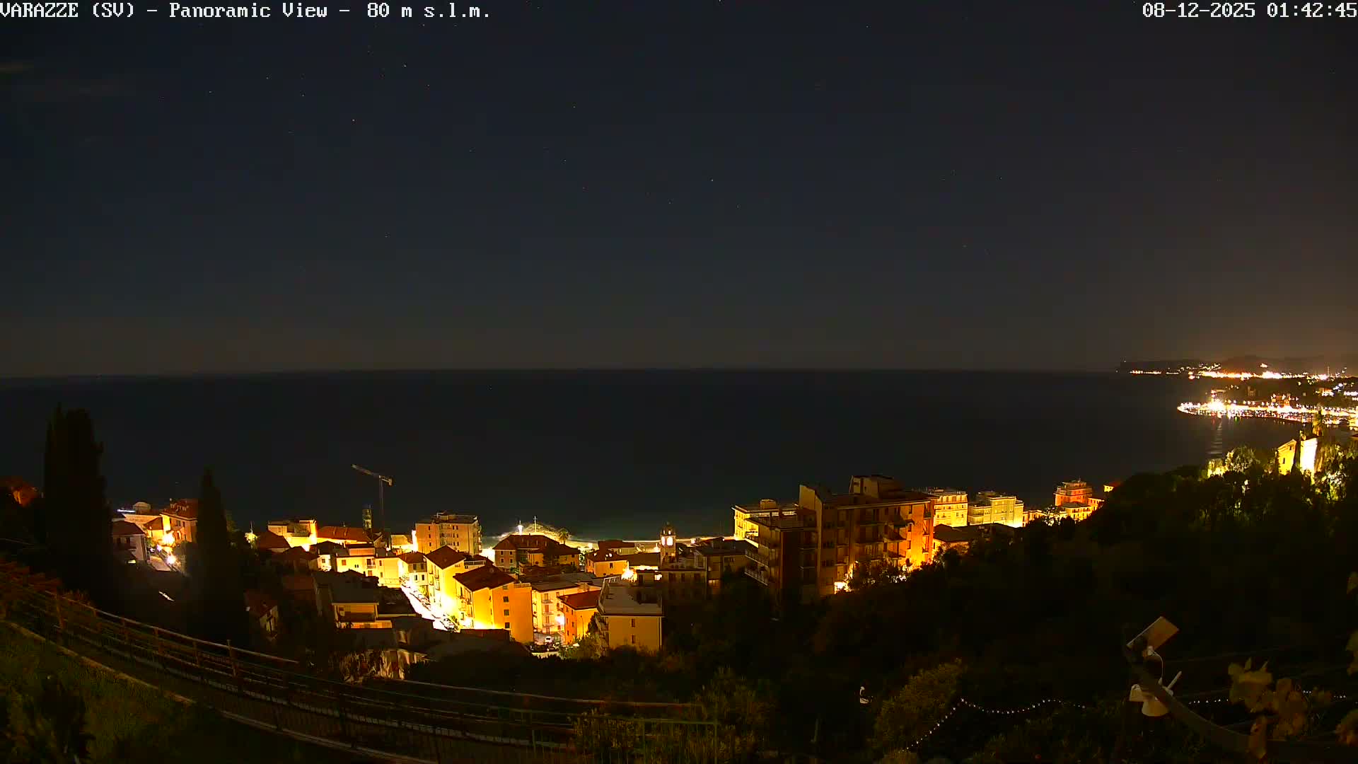 A panoramic night view reveals a brightly lit coastal town nestled along the calm sea, with a long illuminated coastline stretching into the distance under a clear, star-filled sky.
