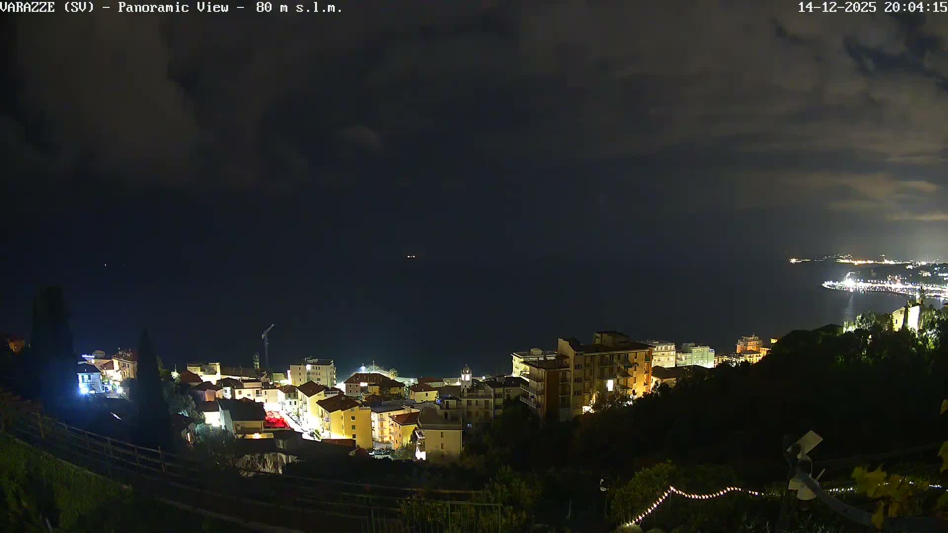 A panoramic night view reveals a brightly lit coastal town nestled along the calm sea, with a long illuminated coastline stretching into the distance under a clear, star-filled sky.