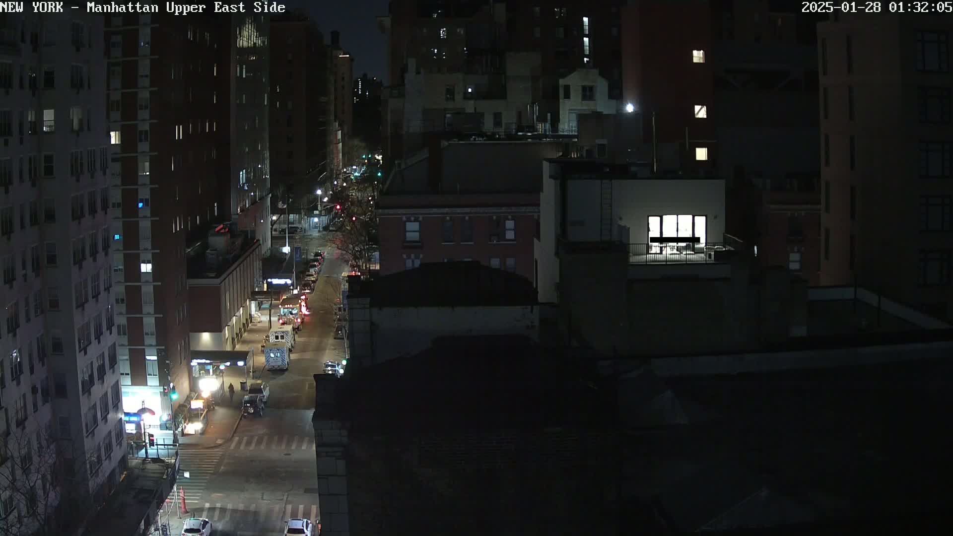 A nighttime aerial view of a city street shows sparsely trafficked roads lined with tall buildings under a clear, dark sky.