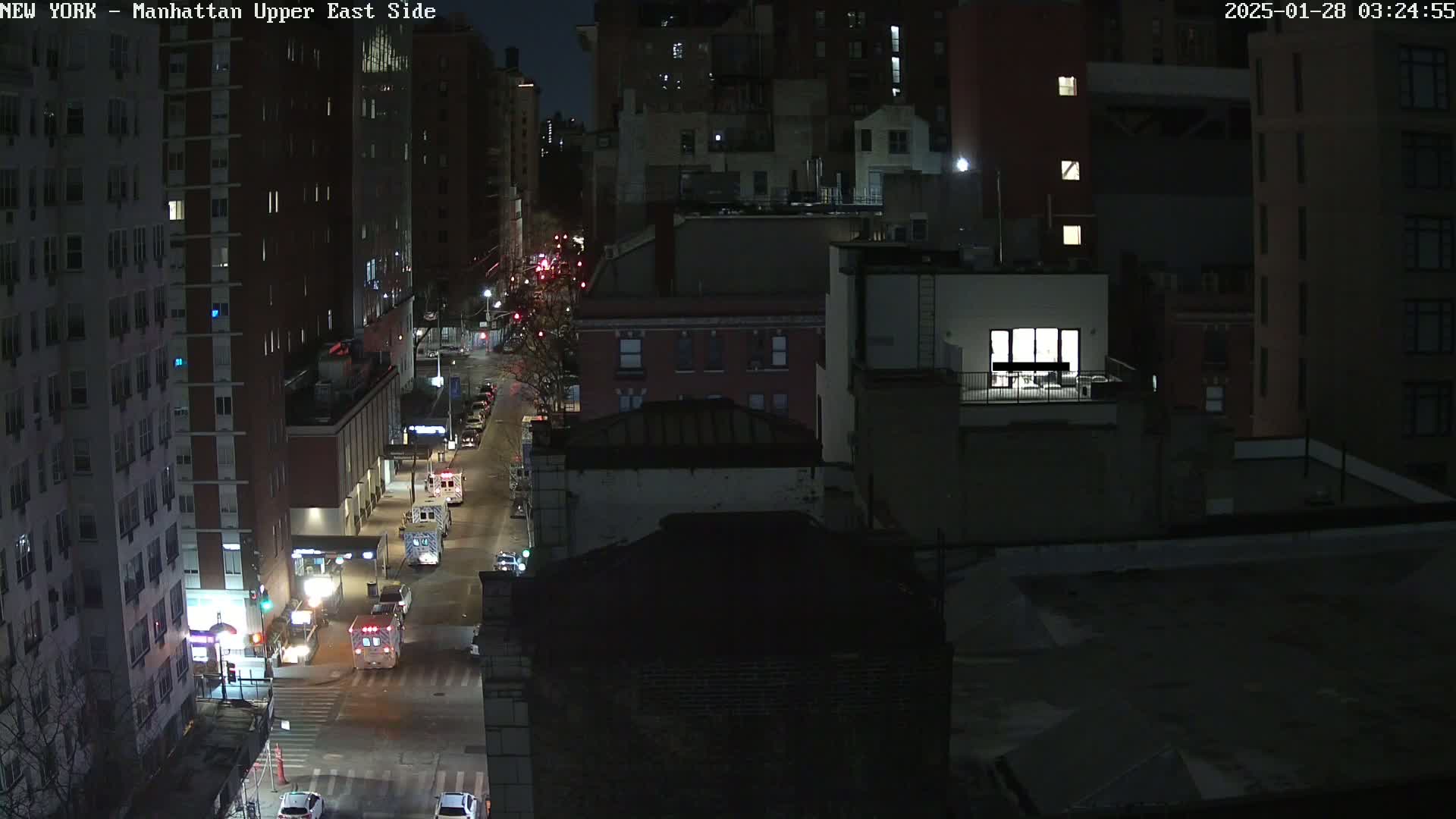 A nighttime aerial view of a city street with several buildings and a few vehicles, including emergency vehicles, under dark, clear skies.