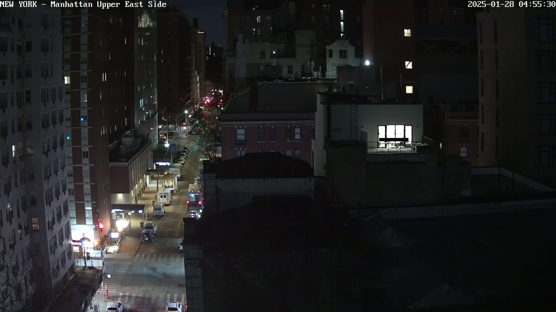 A nighttime aerial view of a city street with several buildings, some vehicles, and a brightly lit apartment balcony.