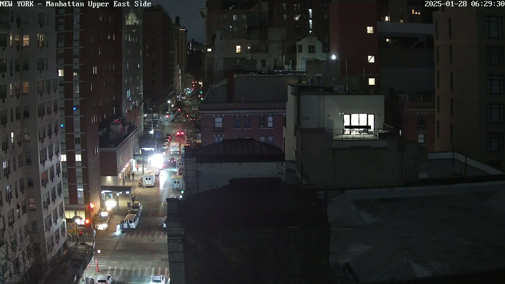 A nighttime aerial view of a city street shows several buildings and vehicles, with some lights visible on the street and in buildings.