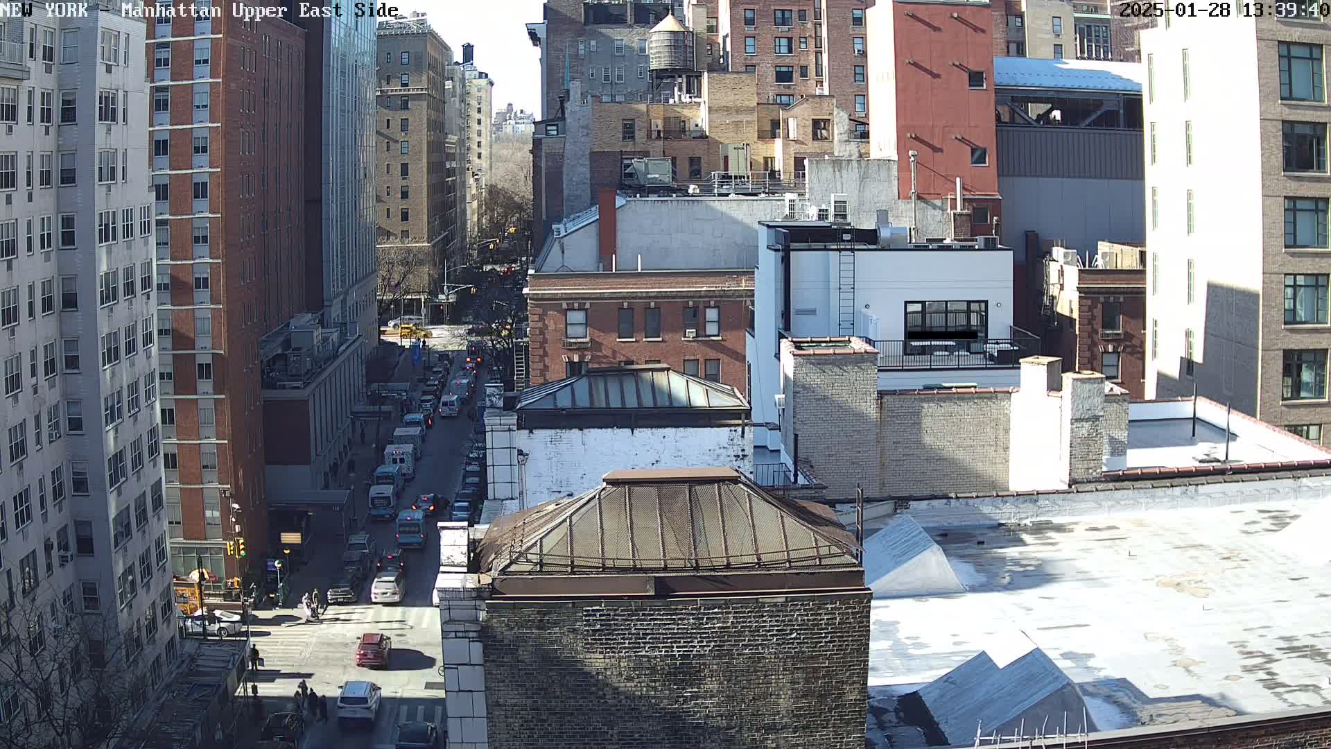 A sunny day view of a city street with traffic and tall buildings, showing rooftops and a water tower.