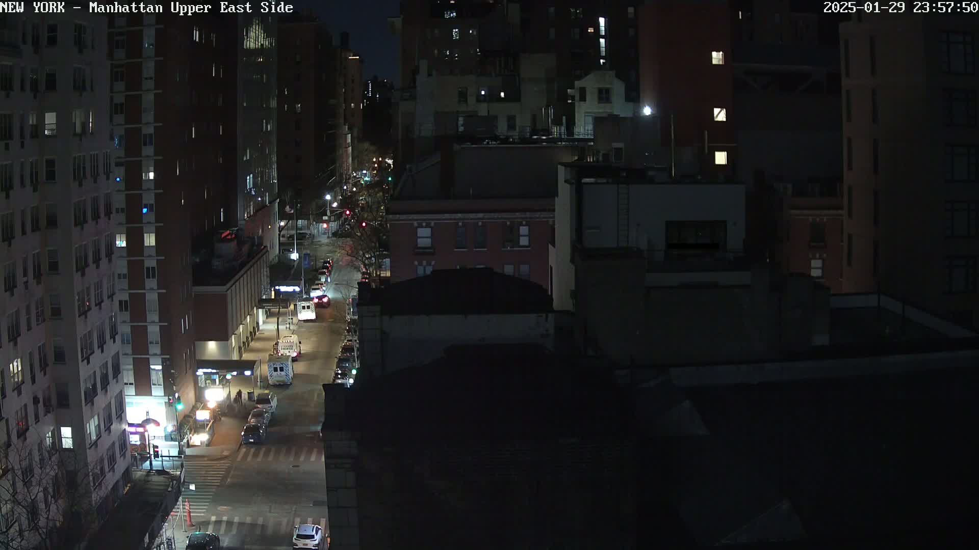 A nighttime, high-angle view of a city street with several cars and buildings, mostly dark except for some illuminated windows.