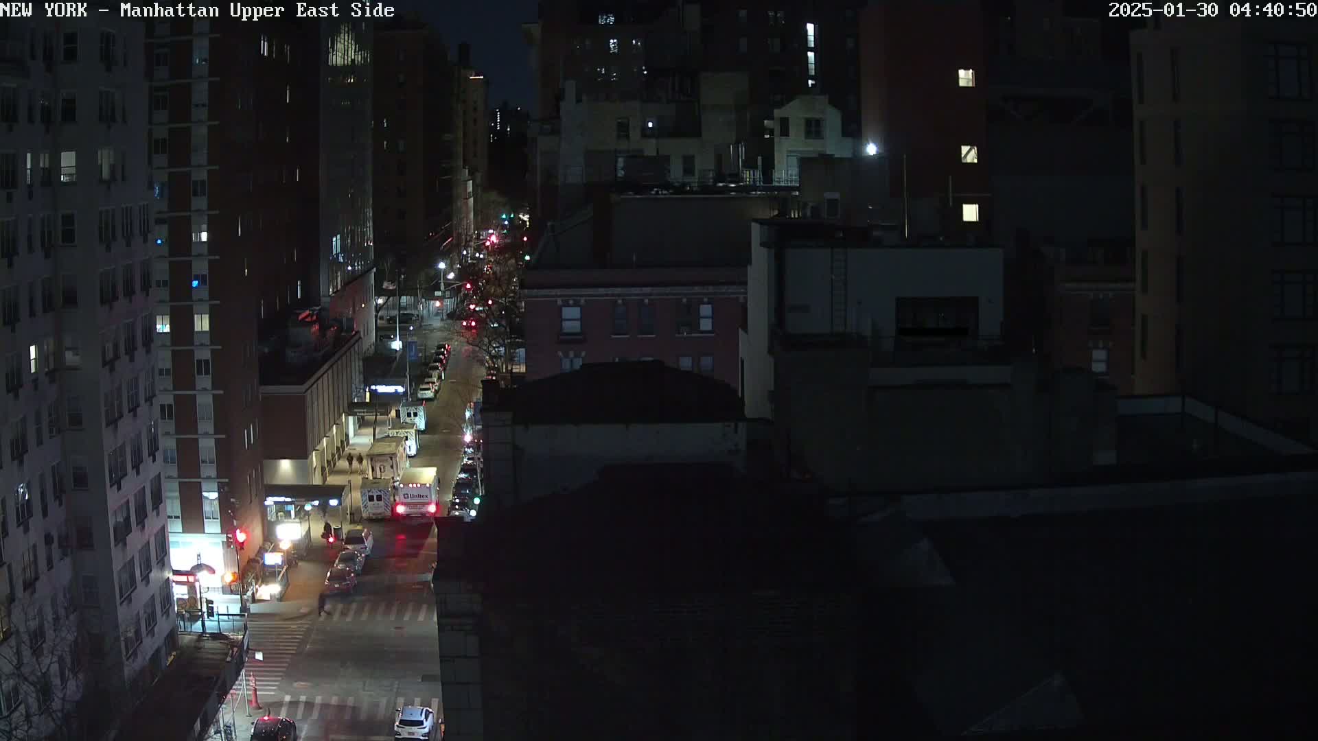 A nighttime, high-angle view of a city street with several buildings and vehicles, under dark conditions.
