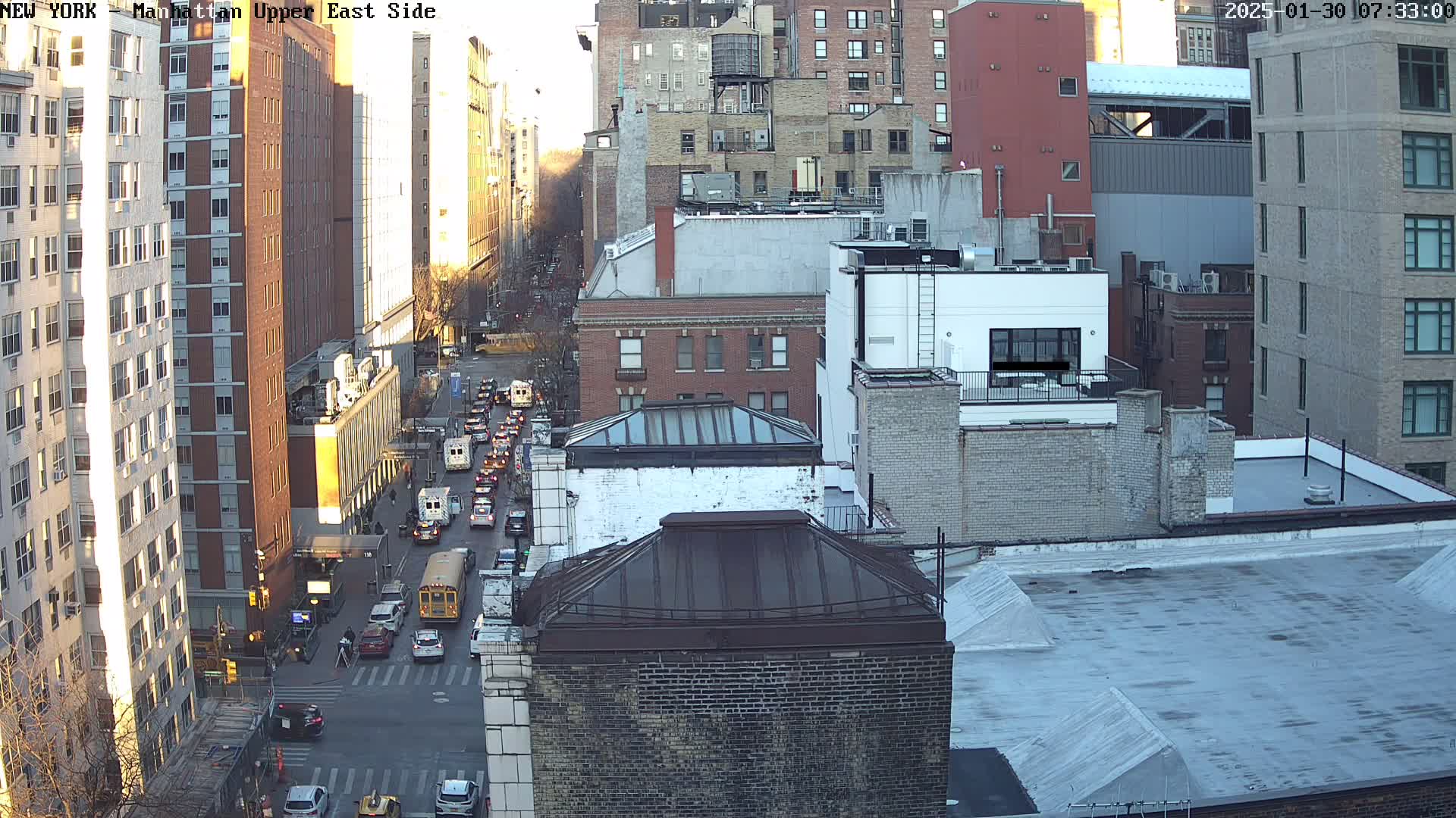 An aerial view of a city street with buildings on either side and a line of slow-moving traffic under a partly sunny sky.