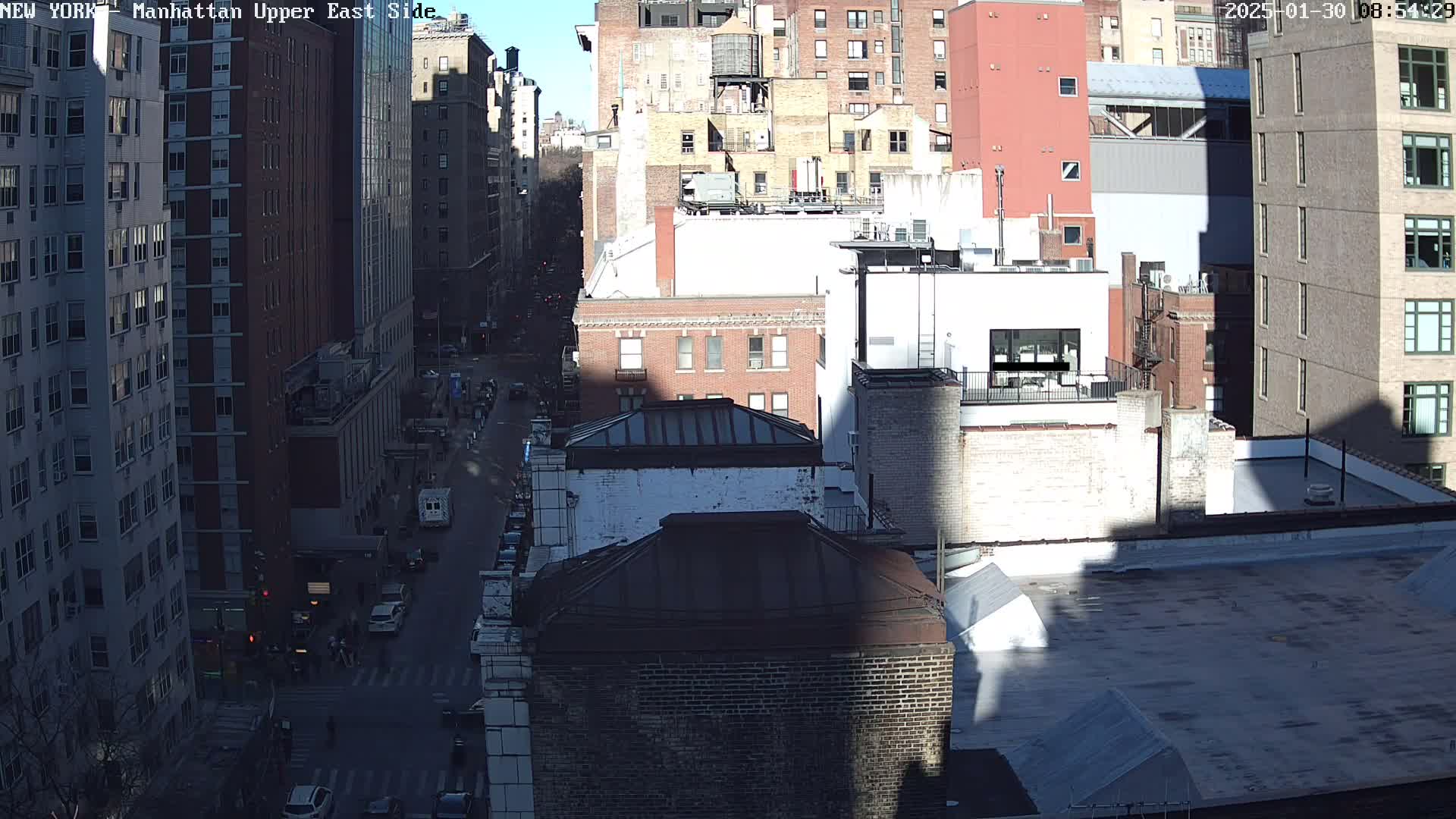 A sunny city street scene shows a bird's-eye view of several multi-story brick buildings, with cars on the street below.