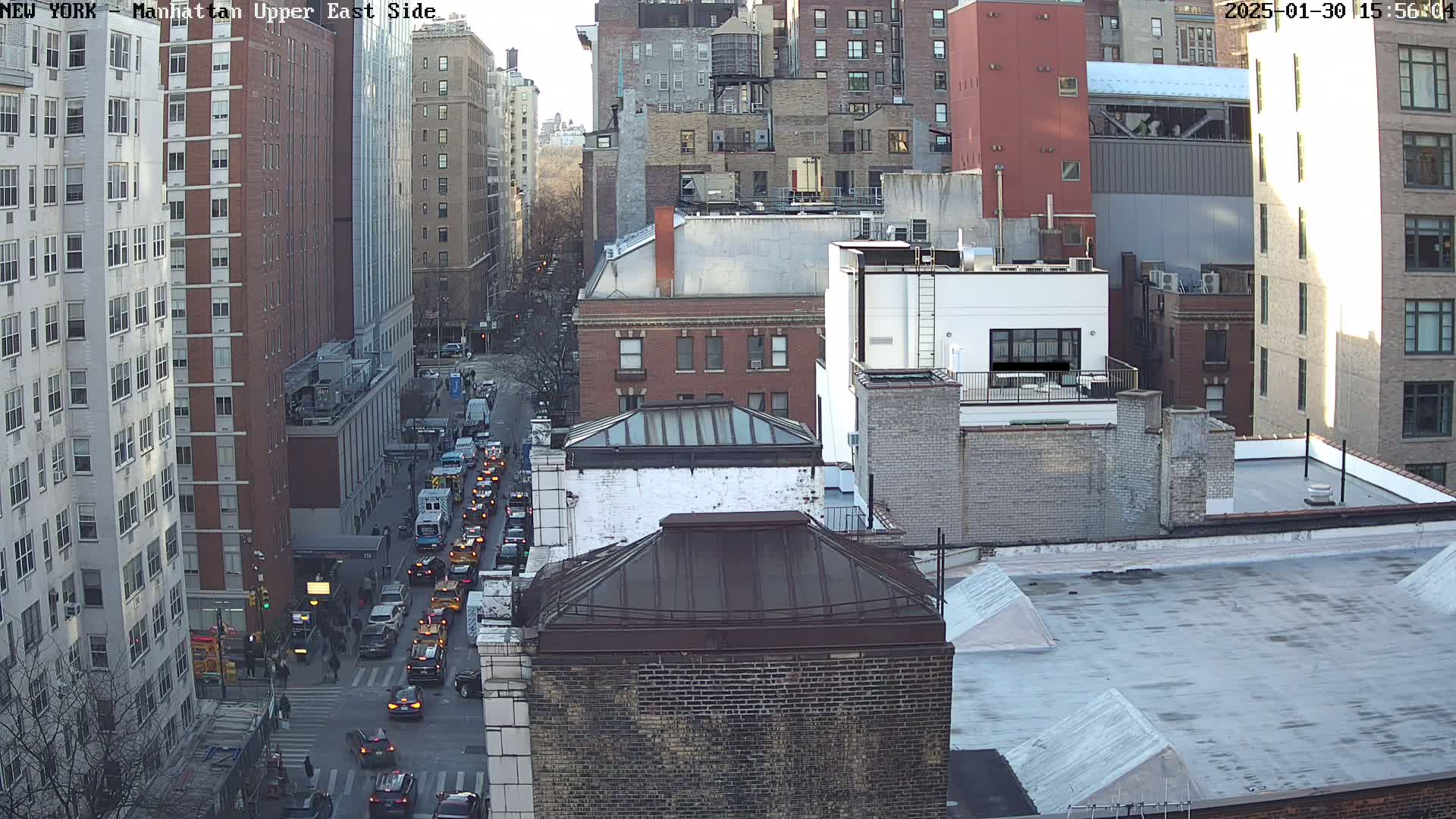 A city street scene on a partly sunny day shows numerous brick buildings and a congested street with cars and buses.