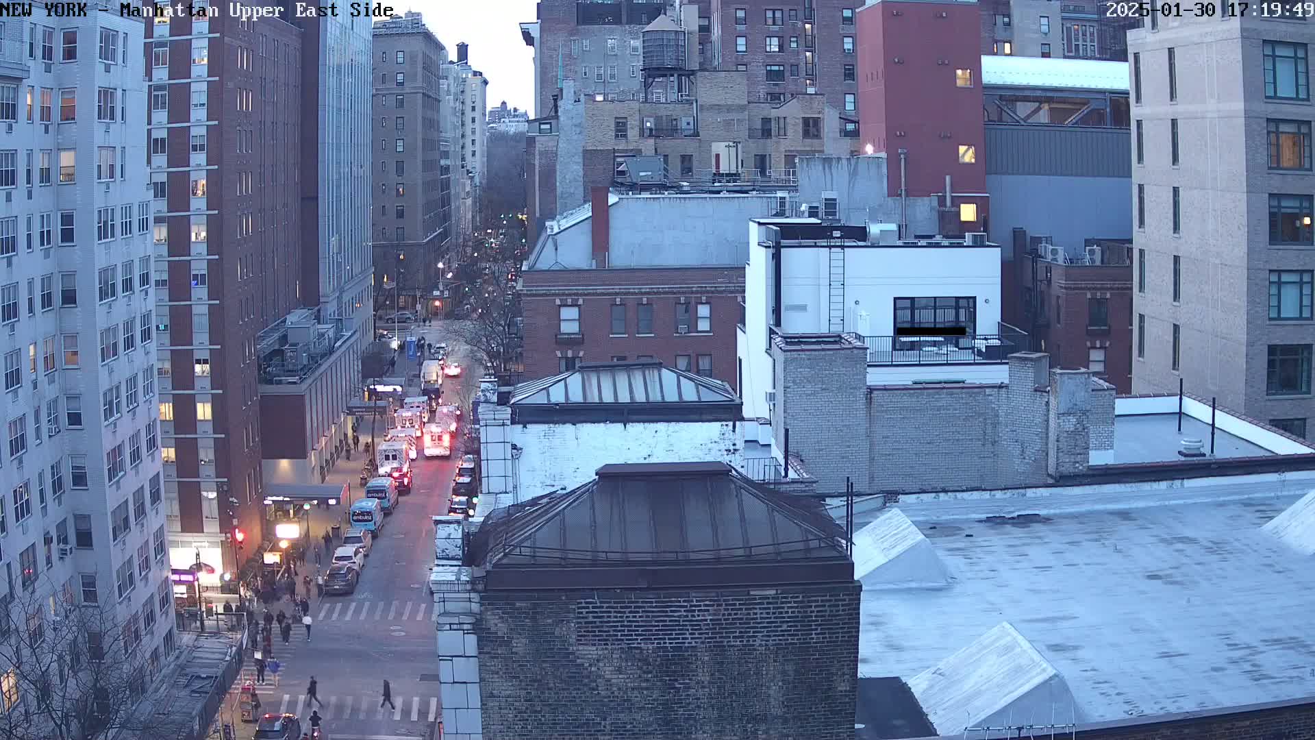 A high-angle, twilight view of a city street with several vehicles, pedestrians, and multi-story buildings.