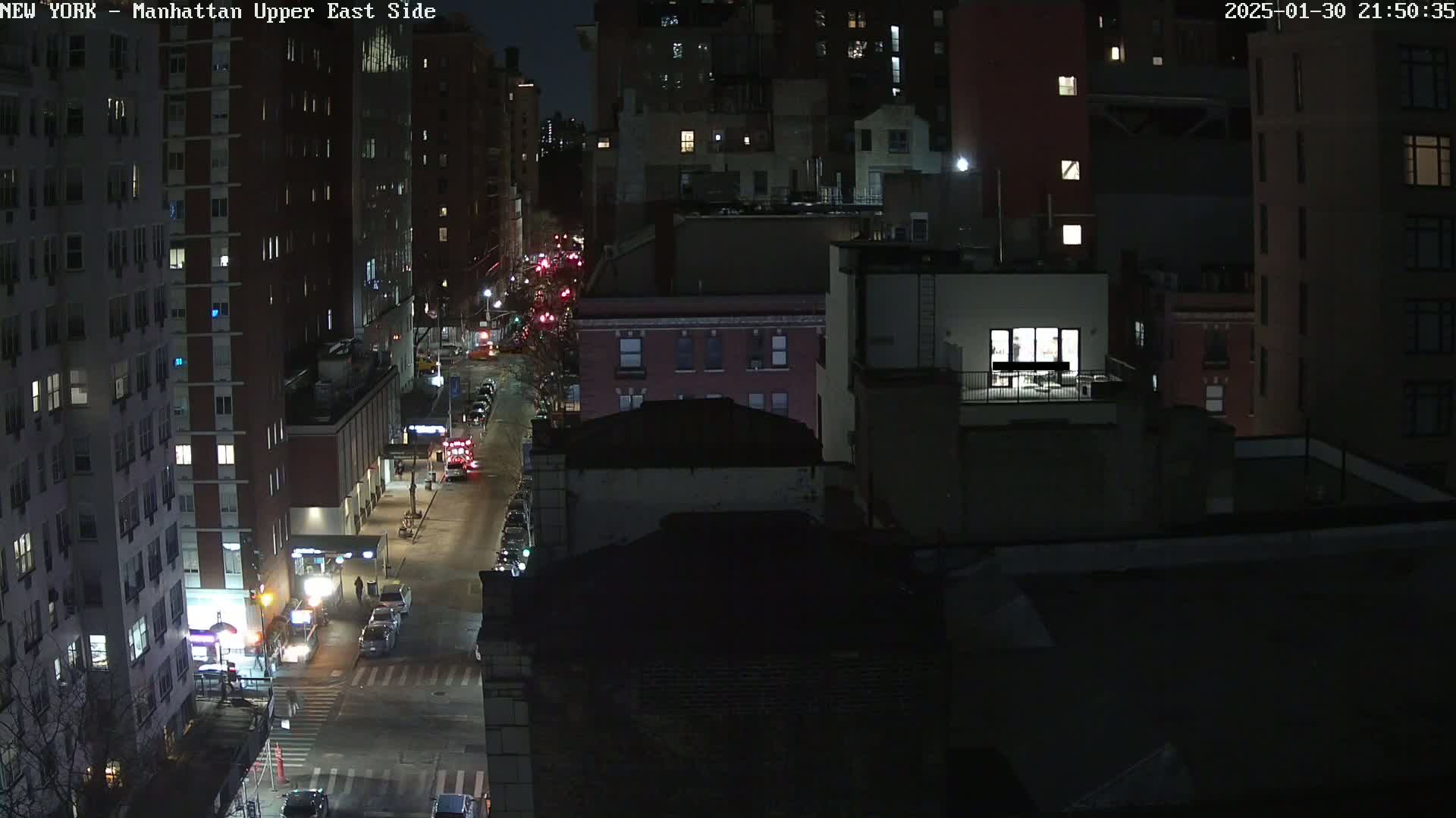 A nighttime, high-angle view of a city street with parked cars and buildings, some with illuminated windows.
