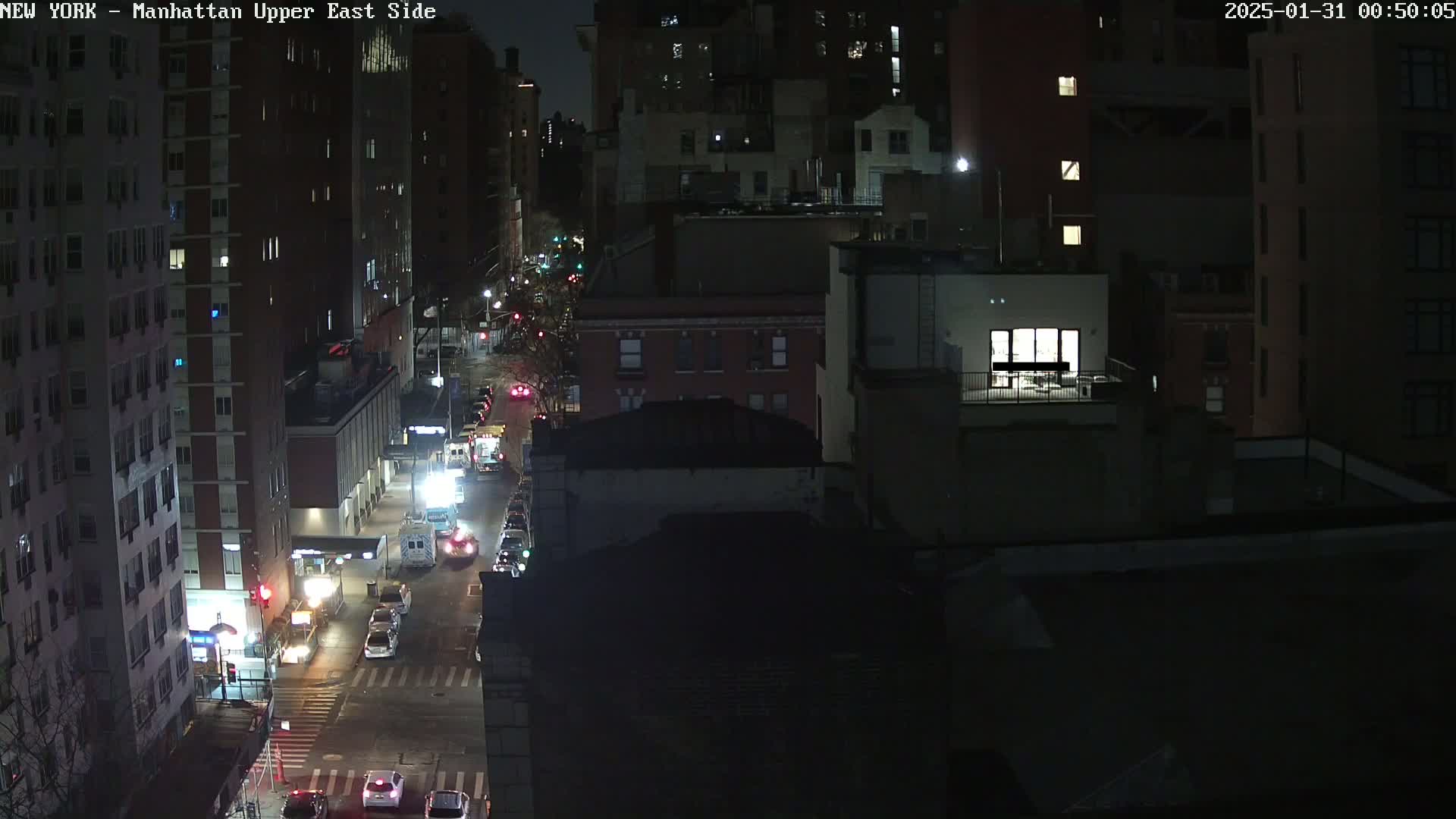 A nighttime aerial view of a city street with several cars and buildings, under dark conditions.