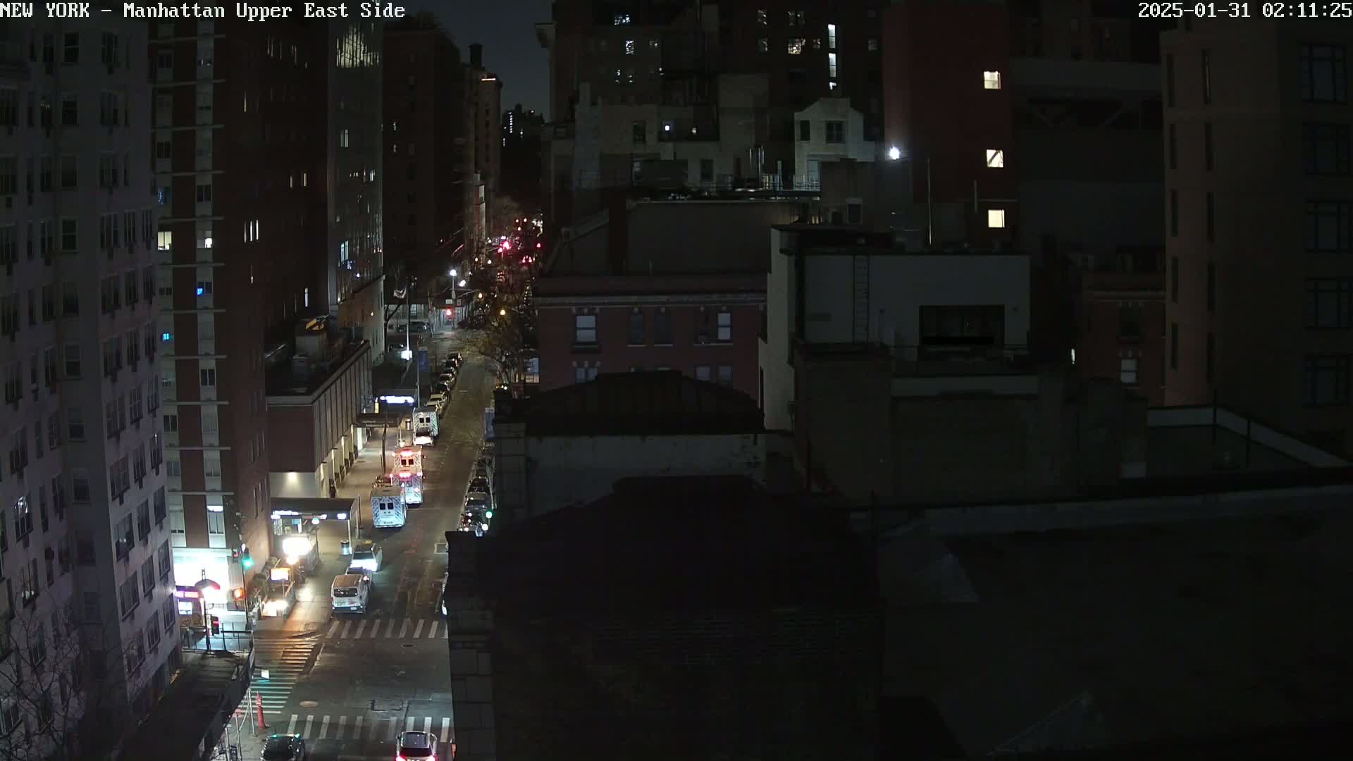 A nighttime aerial view of a city street with several cars and buildings, under dark conditions.