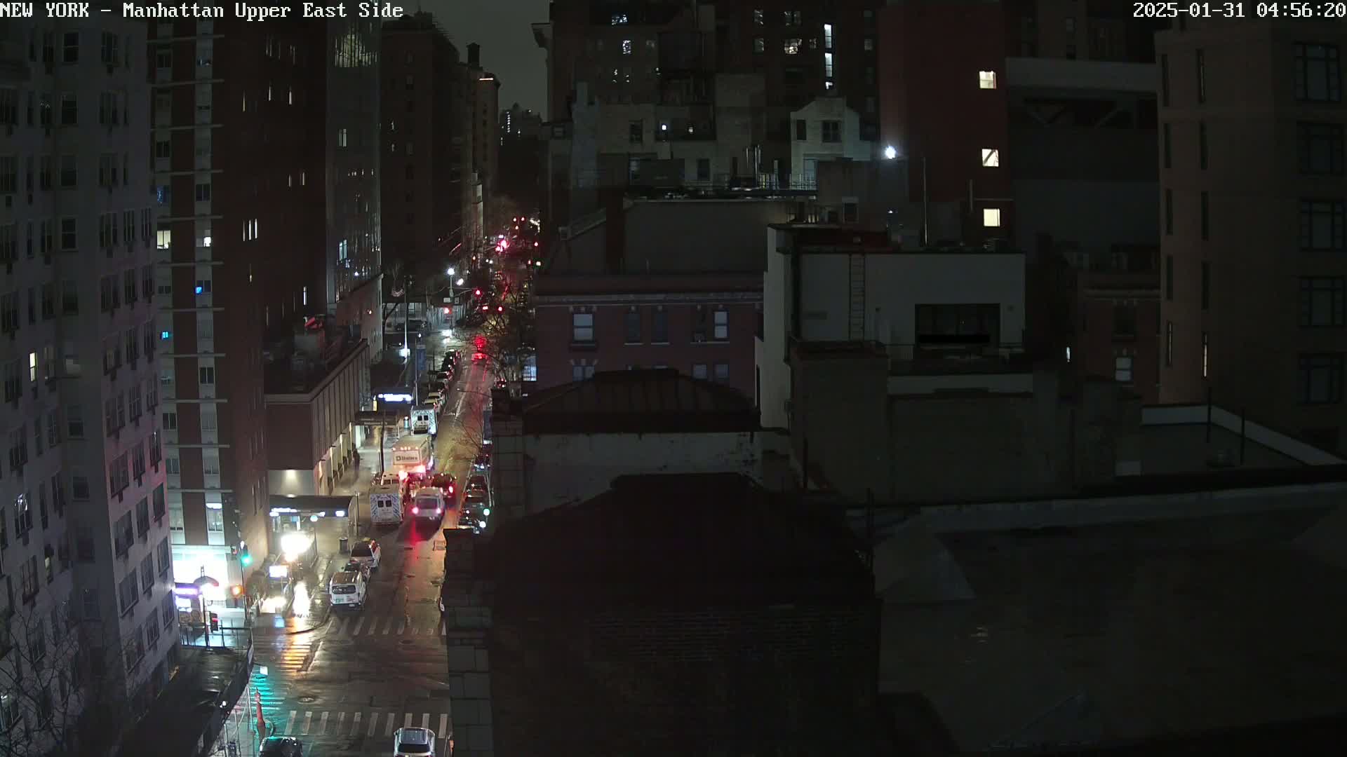 A nighttime, wet street scene shows several vehicles traveling on a city street lined with tall buildings.