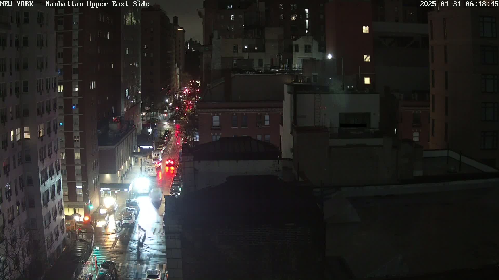 A nighttime, rain-slicked city street scene shows a view of several buildings and traffic moving along a wet road.