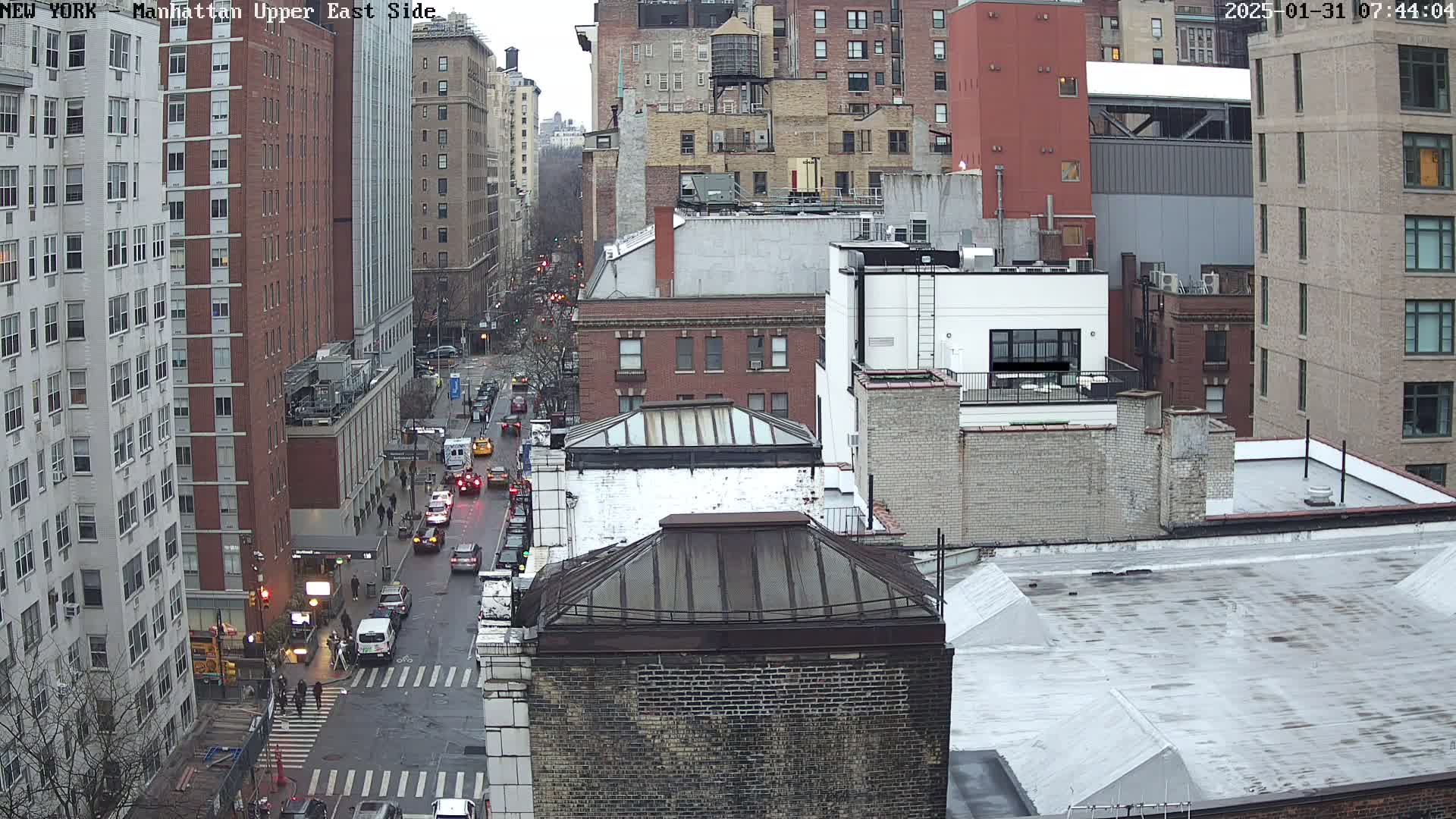 A high-angle, long shot of a city street with several multi-story buildings under an overcast sky, showing some light rain on the street and buildings.