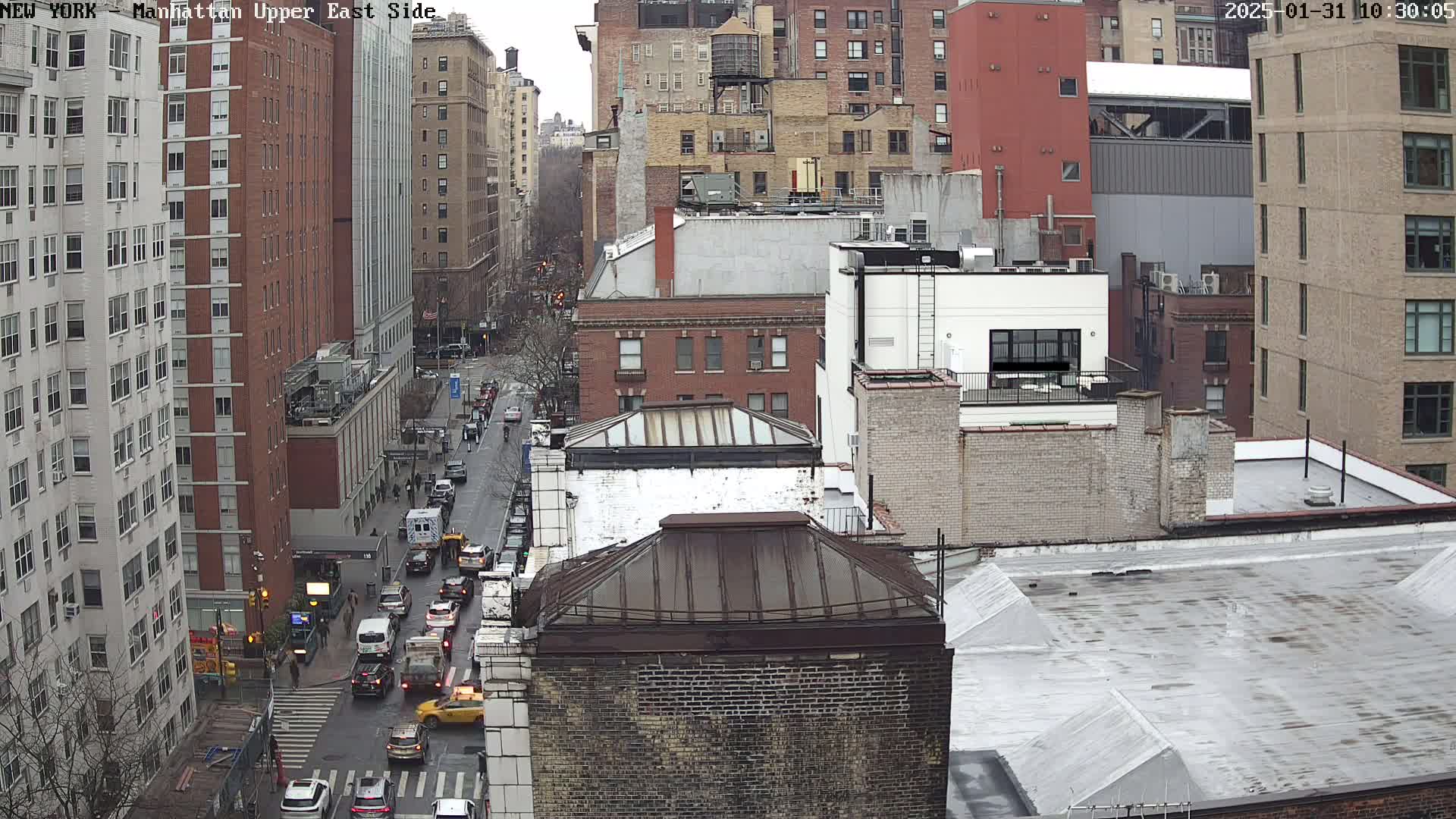 A high-angle, long shot of a city street with multiple buildings, some of which are brick, and cars driving on a wet street during an overcast day.