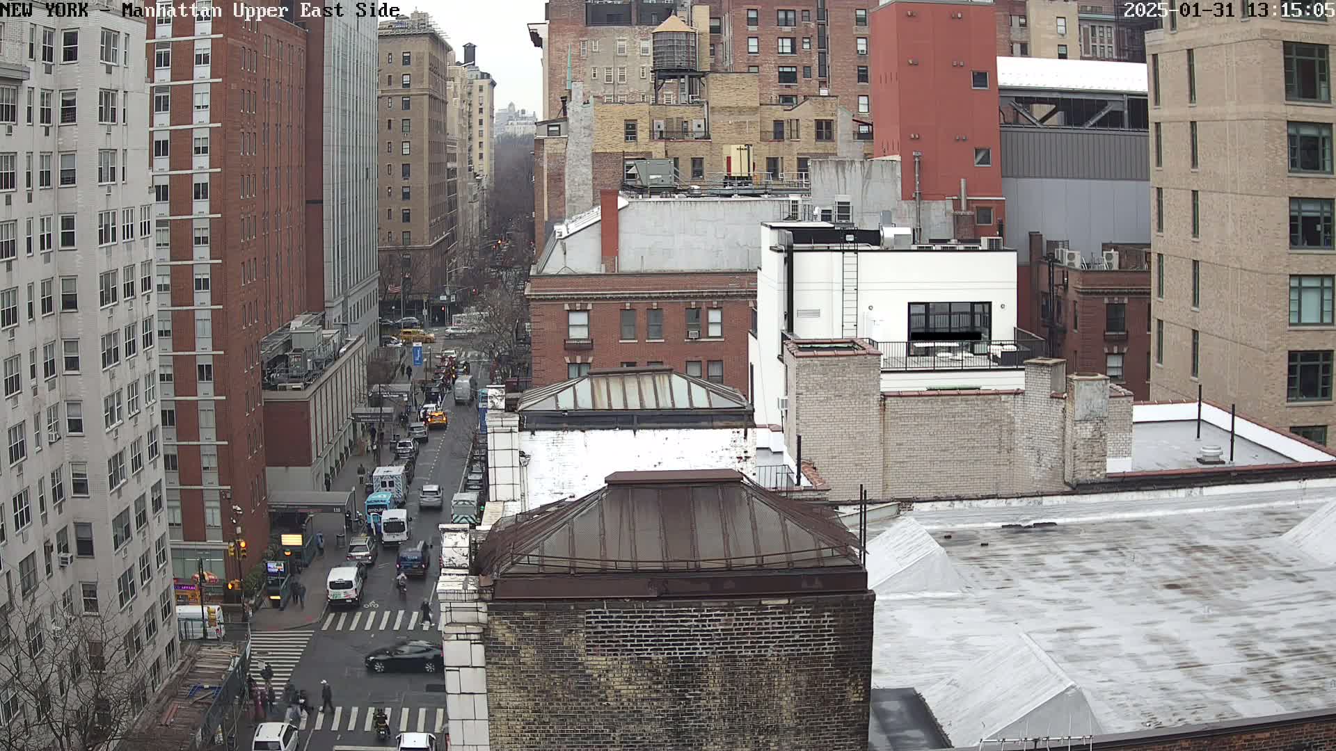 A high-angle view of a city street scene on an overcast day, showing several multi-story buildings, vehicles on the street, and pedestrians at a crosswalk.