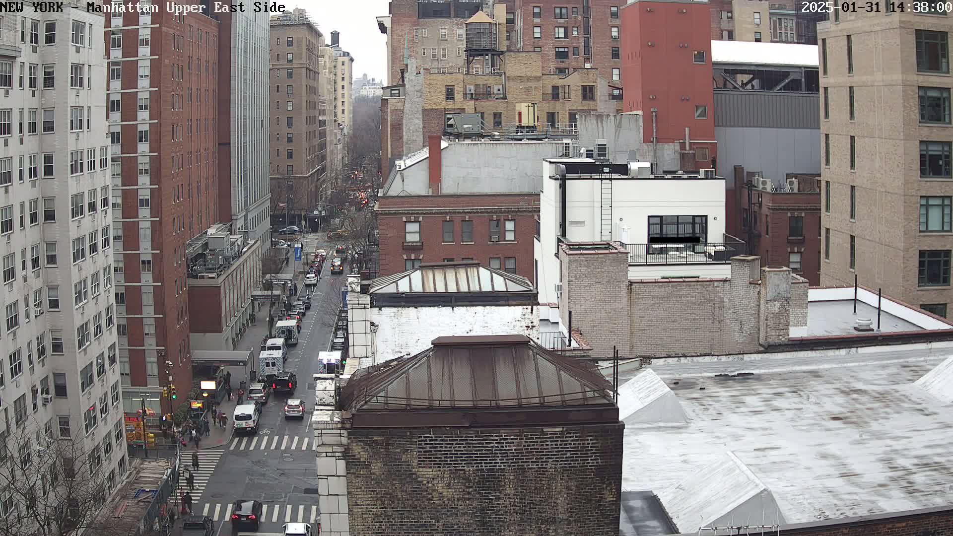 A high-angle view of a city street scene on an overcast day, showing multiple buildings, vehicles, and pedestrians at a street intersection.