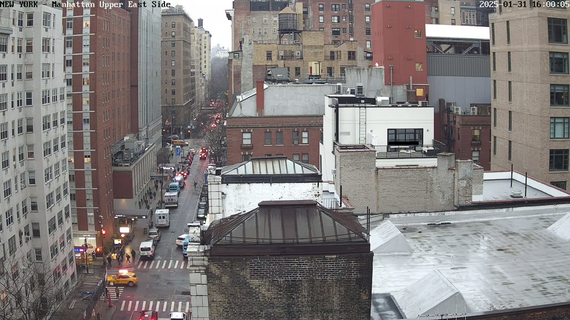 A high-angle view of a city street, showing several multi-story buildings, vehicles, and pedestrians on a cloudy day.