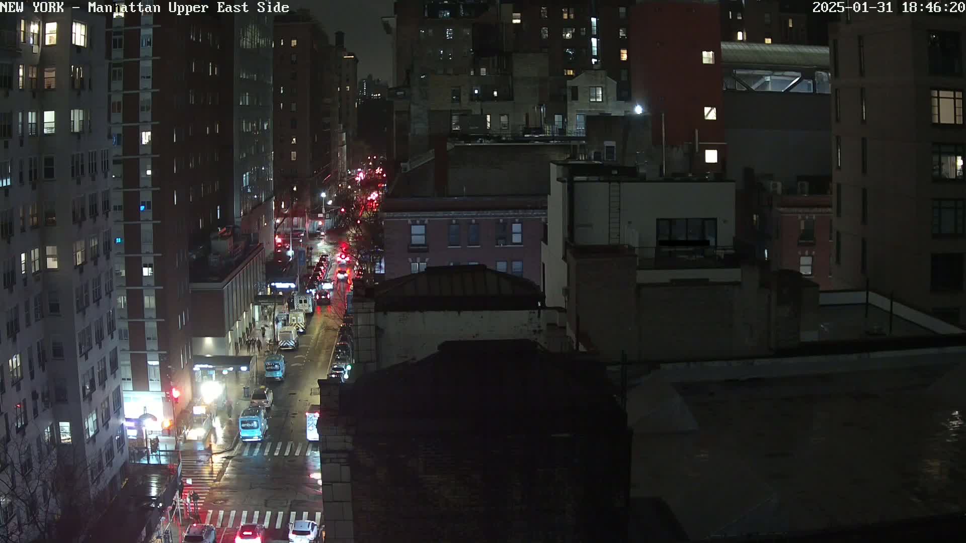A nighttime, wet city street scene shows traffic moving along a road between tall buildings under dark, overcast skies.
