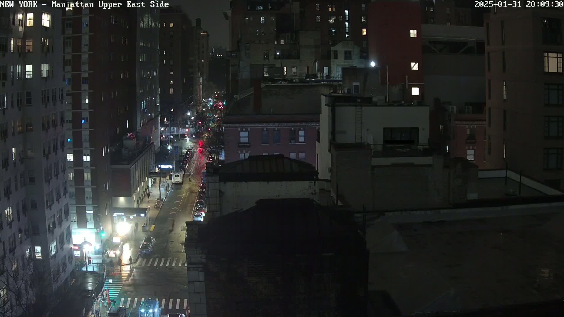 A nighttime, rain-slicked city street scene shows several tall buildings and a few vehicles on a roadway, viewed from an elevated position.