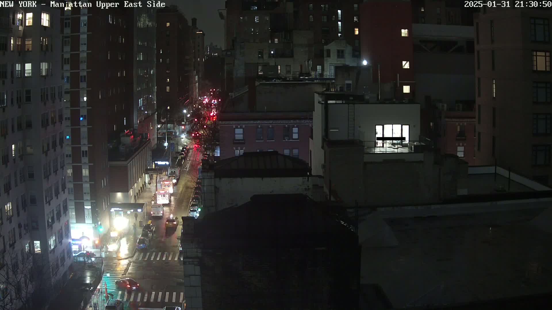 A nighttime, wet city street scene shows traffic moving along a road between tall buildings under dim lighting.