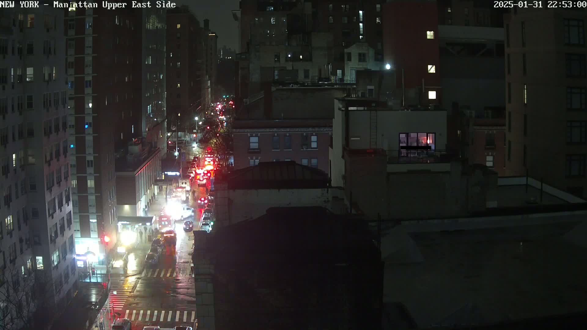 A nighttime, wet city street scene shows congested traffic between tall buildings under dark, cloudy skies.