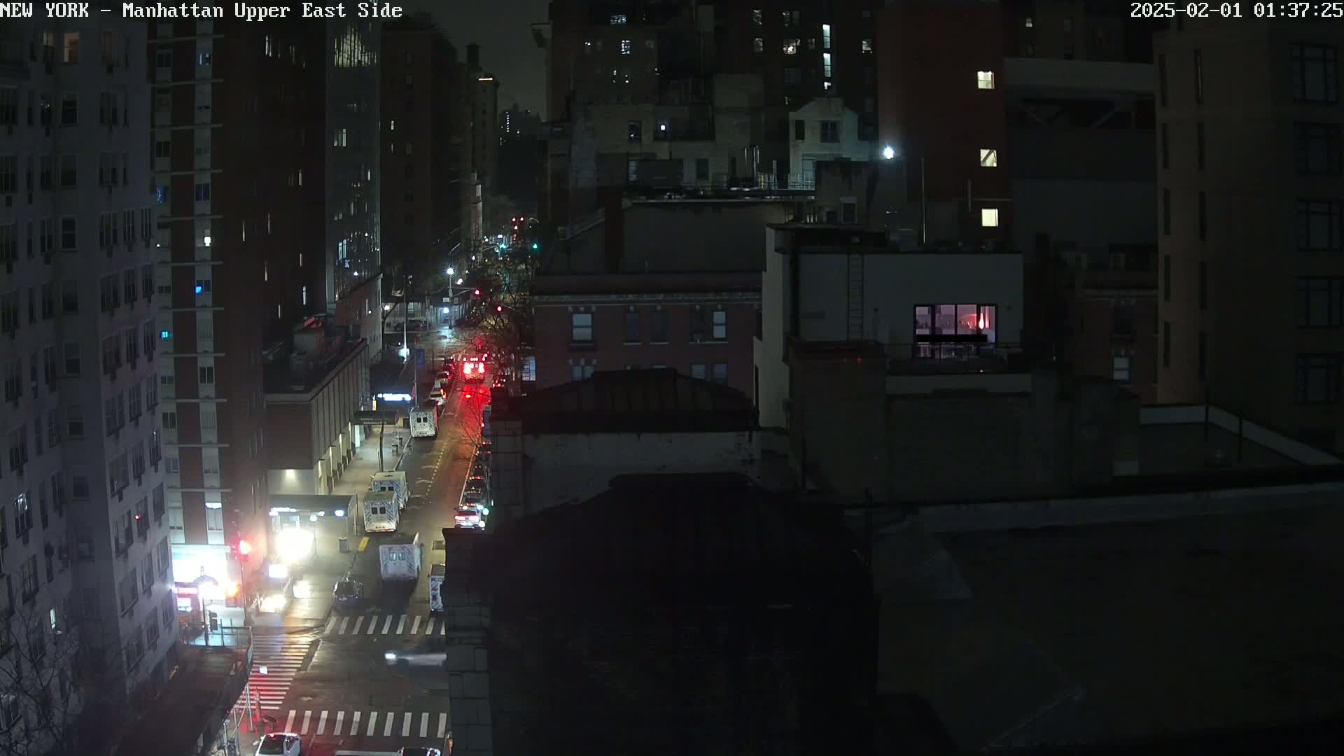 A nighttime aerial view of a city street shows several cars driving on a wet road between tall buildings under dark skies.