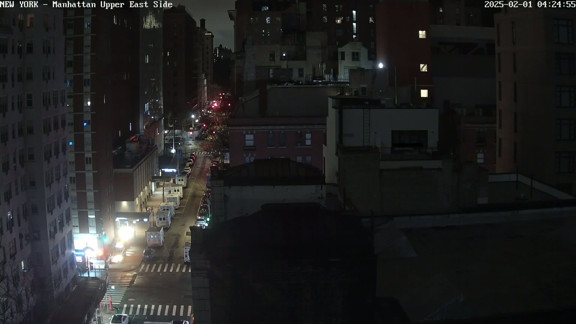 A nighttime, overhead view of a city street with several multi-story buildings, some vehicles parked and driving, and a wet road.