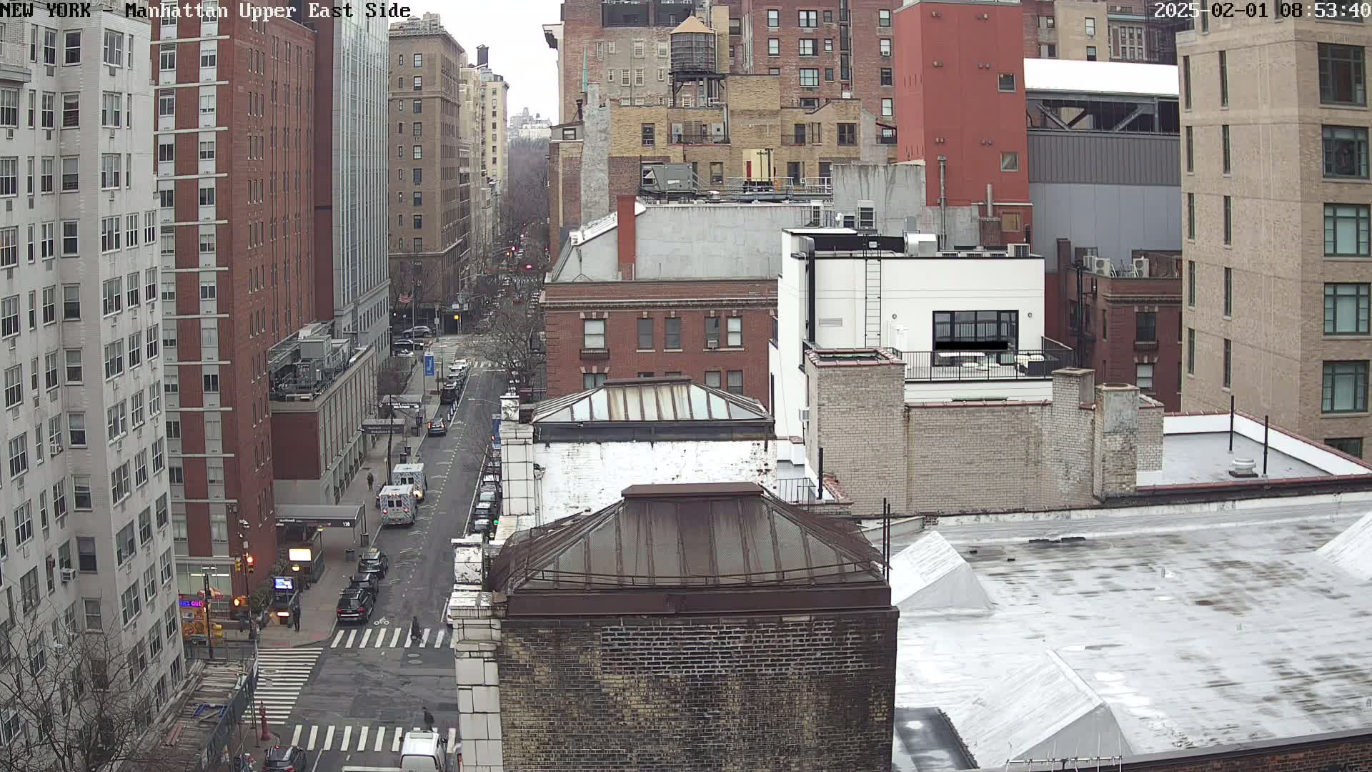 A high-angle, long shot of a city street on an overcast day, showing several multi-story brick buildings and a few vehicles on the street below.