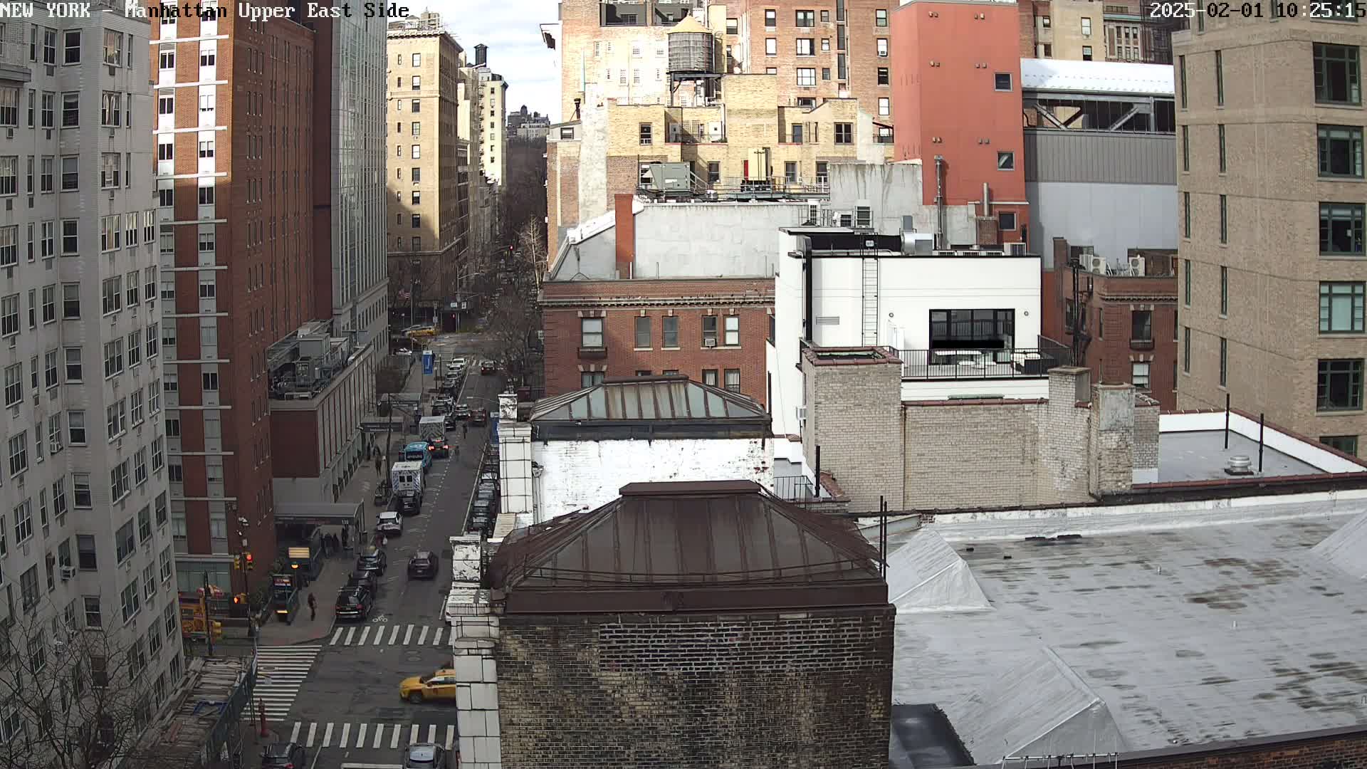 A high-angle view of a city street on a sunny day, showing multiple brick buildings and cars traveling along the road.