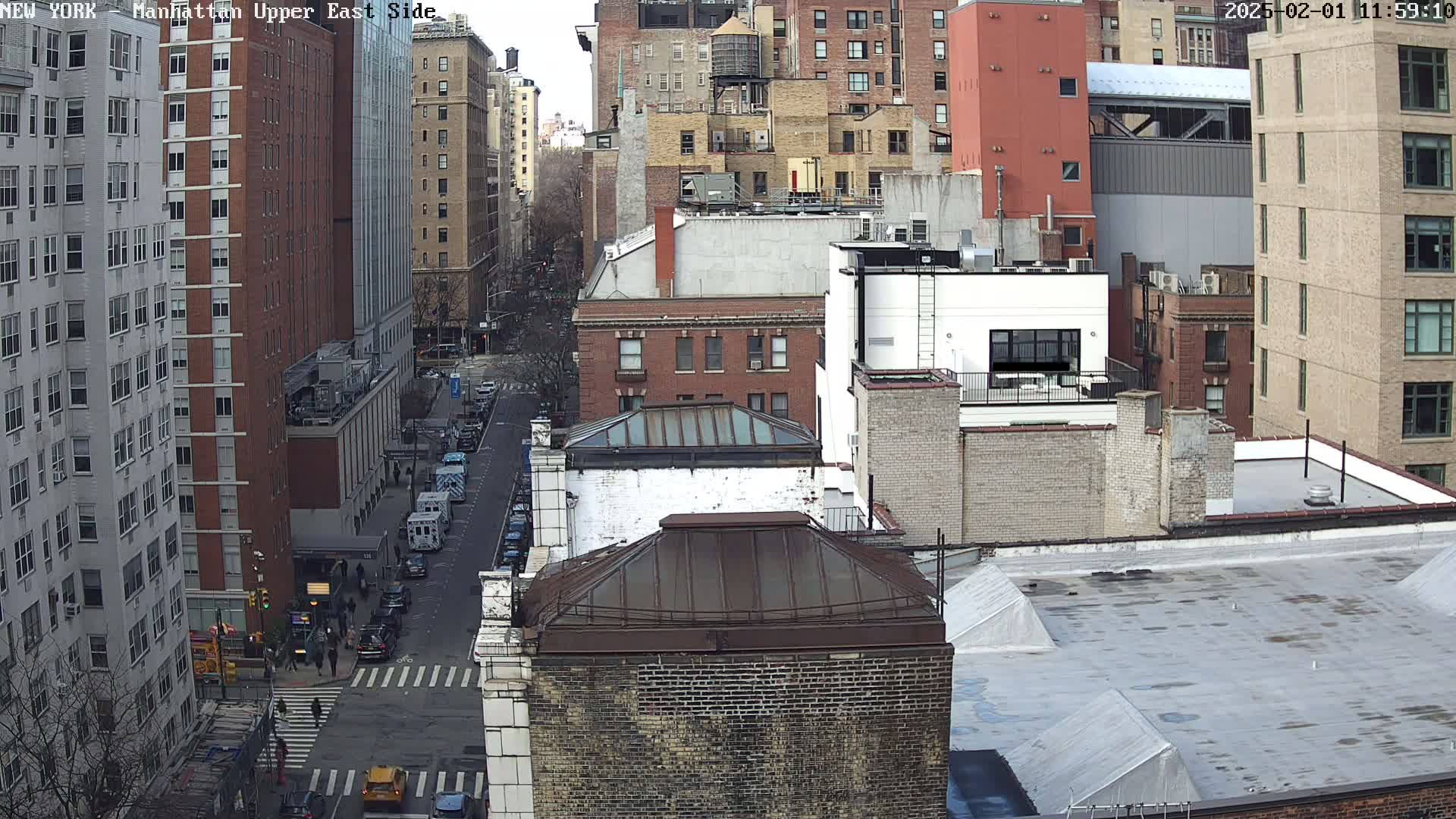 A high-angle view of a city street scene on an overcast day, showing several multi-story brick buildings, a street with cars and pedestrians, and a mix of architectural styles.