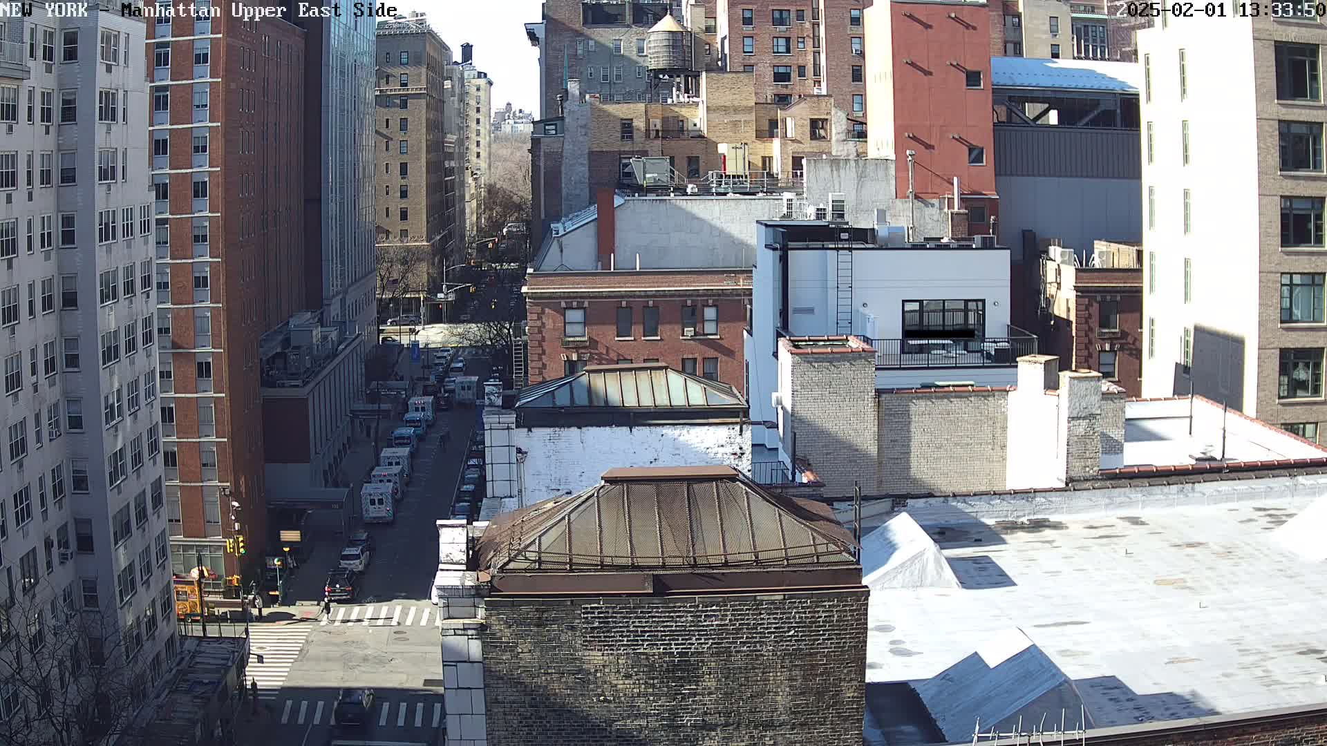 A sunny cityscape view of several multi-story brick buildings, some with rooftop structures, and a street with vehicles, including several emergency vehicles.