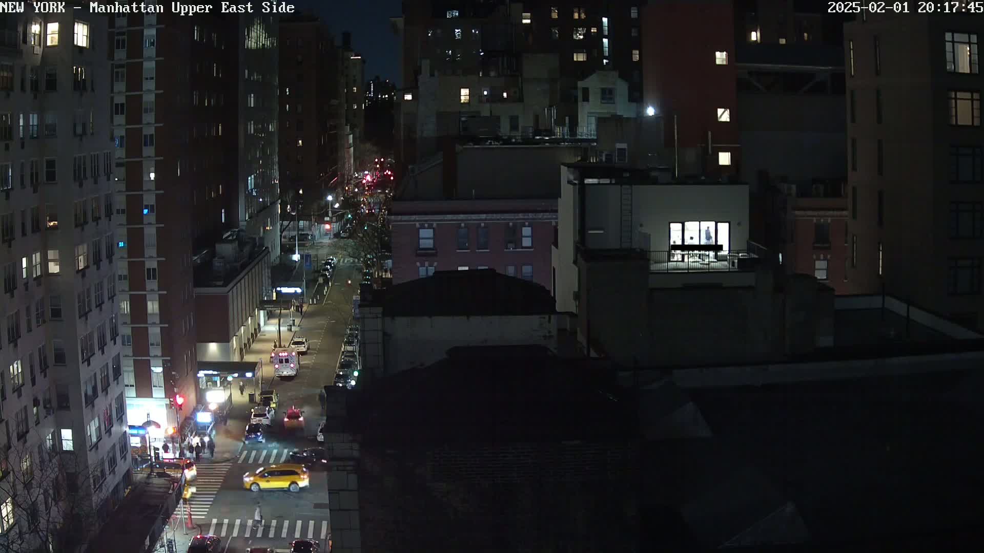 A nighttime aerial view of a city street shows several multi-story buildings, cars, and pedestrians at an intersection.