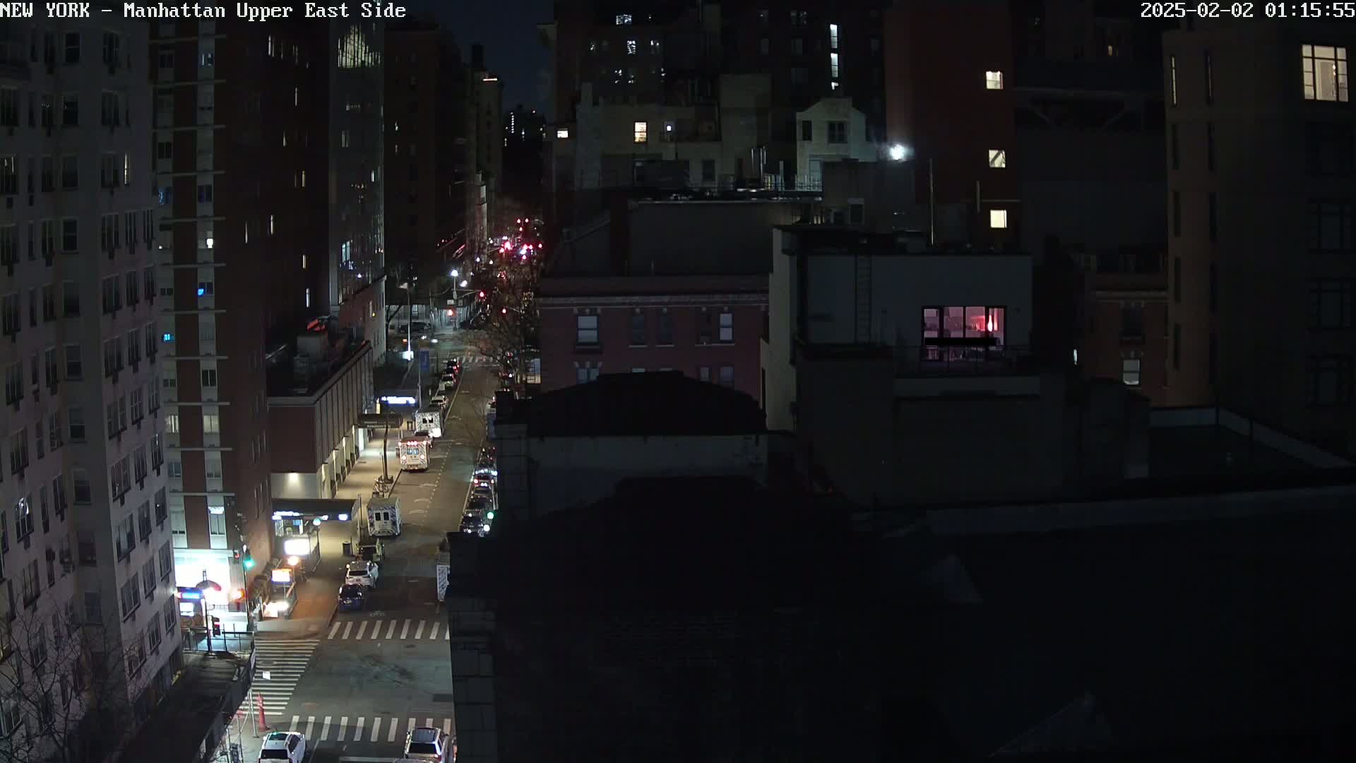 A nighttime, aerial view of a city street with sparsely parked cars and several multi-story buildings.
