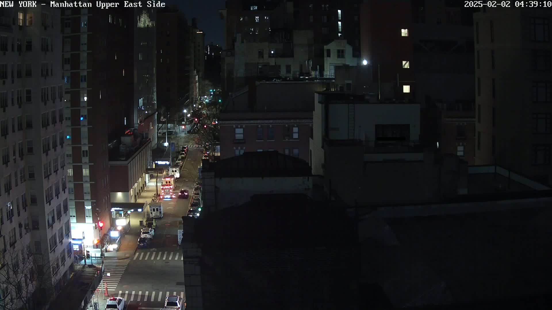 A nighttime aerial view of a city street with several buildings and vehicles, including emergency vehicles.