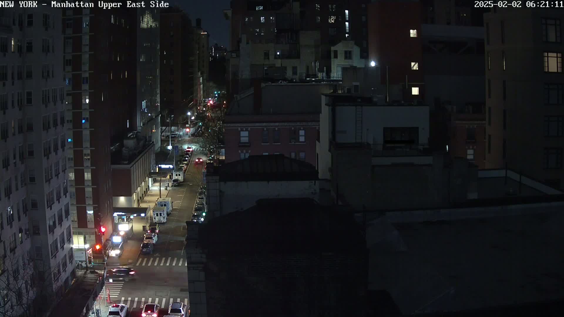 A nighttime aerial view shows a city street with sparse vehicle traffic between tall buildings under a dark sky.