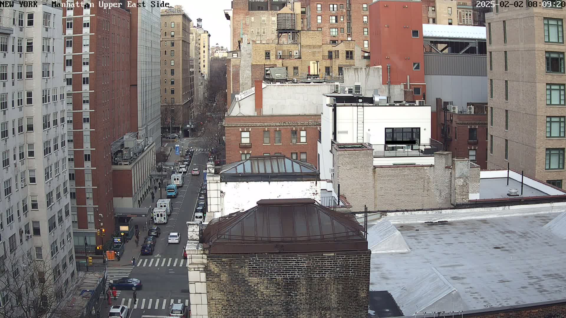 A high-angle, long shot of a city street, with several multi-story buildings on either side, under an overcast sky.