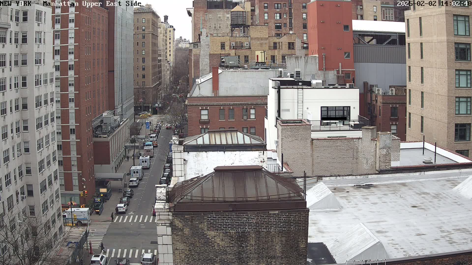 A high-angle view of a city street scene on a cloudy day, showing several multi-story brick buildings, a street with cars, and emergency vehicles near a hospital.