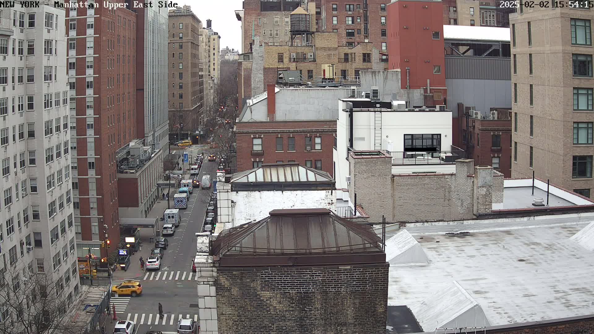 An overcast day view of a city street, showing several tall brick apartment buildings and a few vehicles on the road below.
